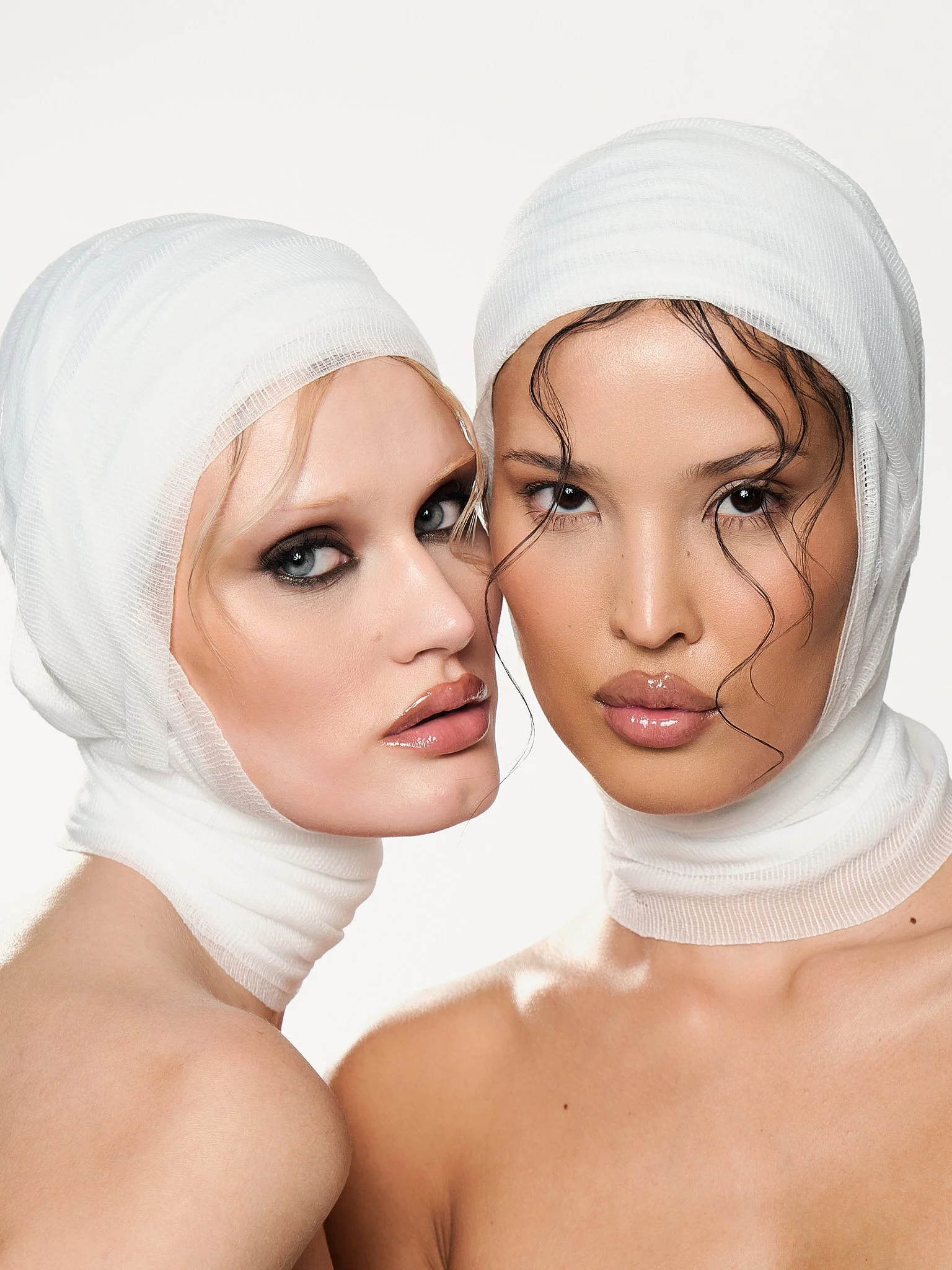 Close-up of two women with light and dark skin tones wearing white head coverings and minimal makeup, posing closely together against a white background.