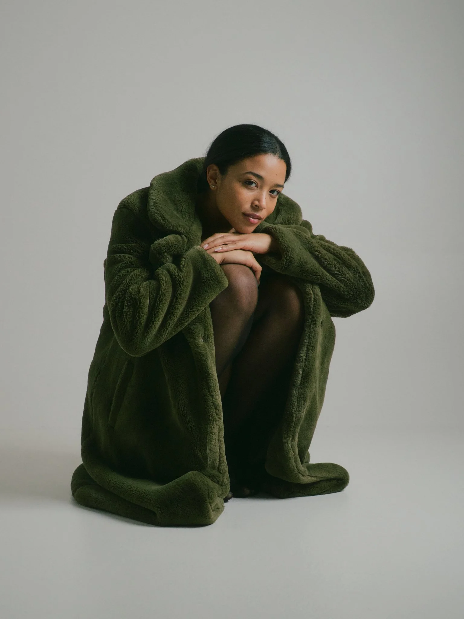 A woman with dark hair and earrings, wearing a green fuzzy coat, sitting on the floor with her knees up and arms resting on her knees, against a plain gray background.