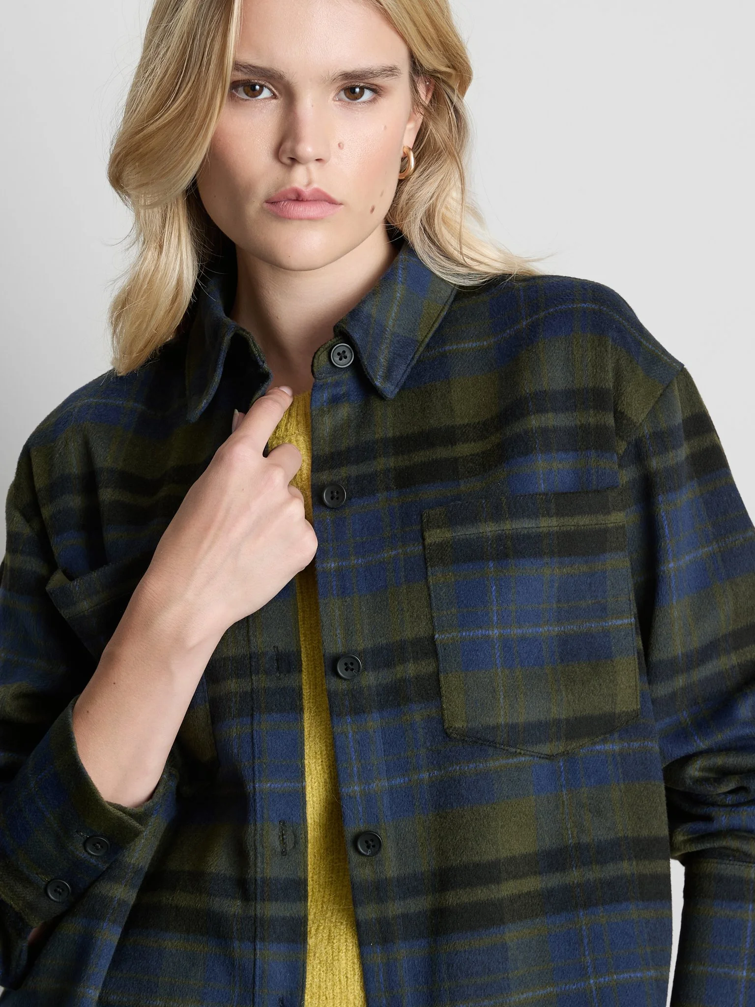 A young woman with blonde hair and brown eyes wearing a dark plaid shirt over a yellow sweater.