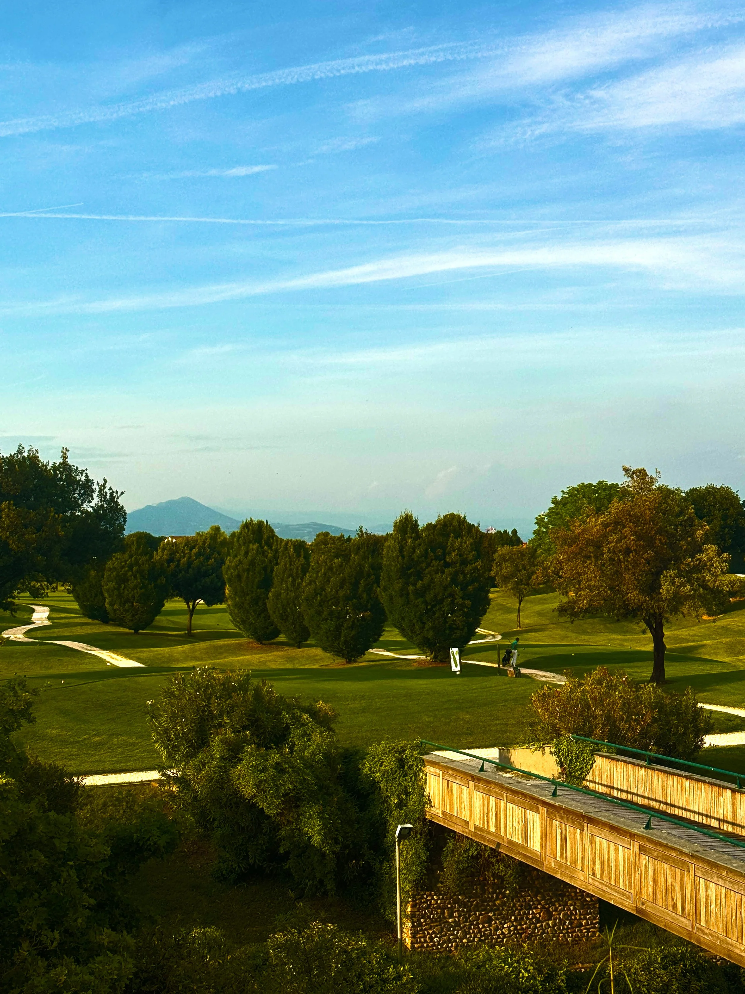 A scenic view of a golf course with lush green grass, trees, and a distant mountain range under a blue sky with wispy clouds.