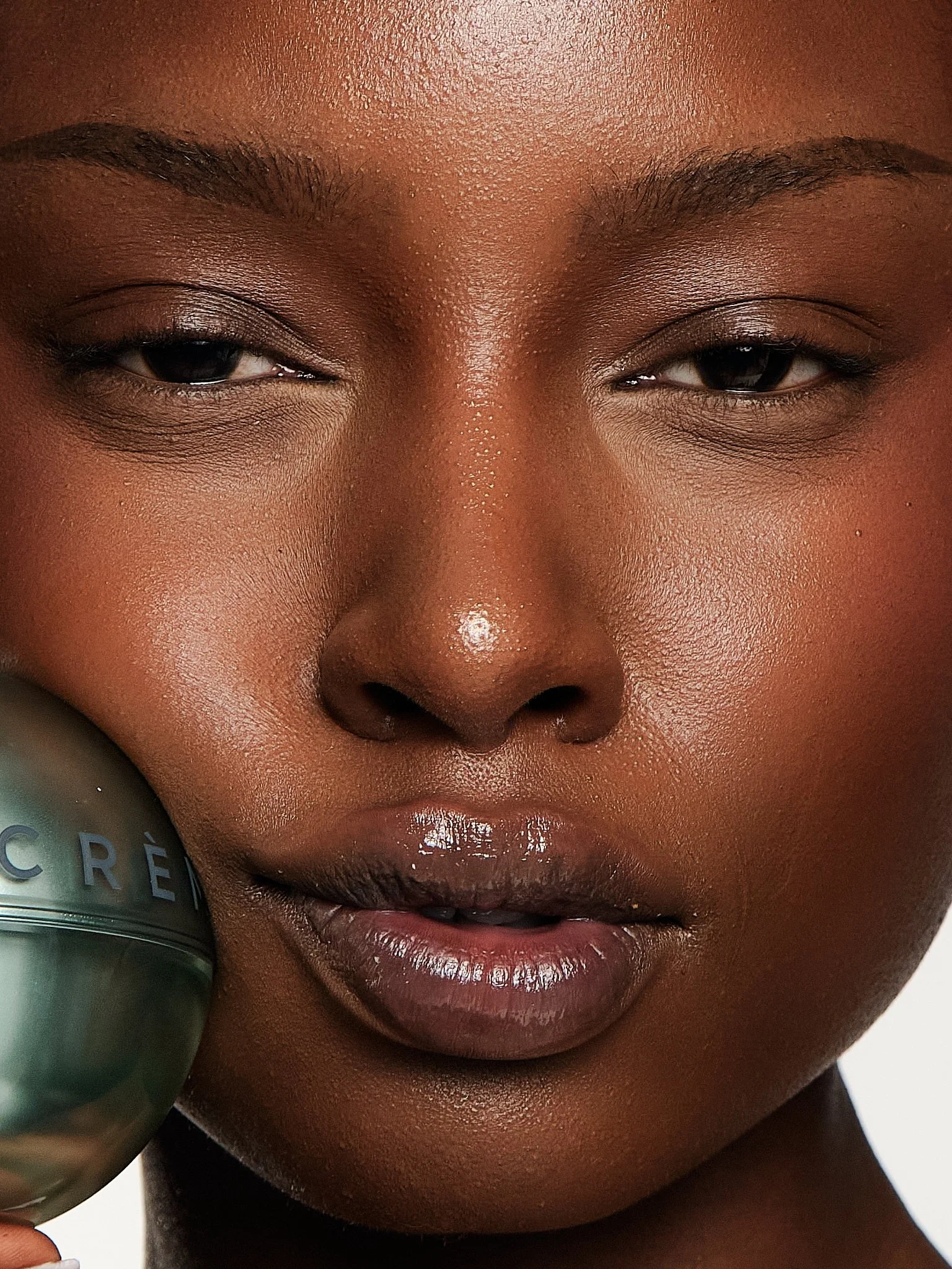 Close-up of a person's face with dark skin, with eyes closed and lips slightly parted, holding a grey container with the brand name 'crème' near the cheek.