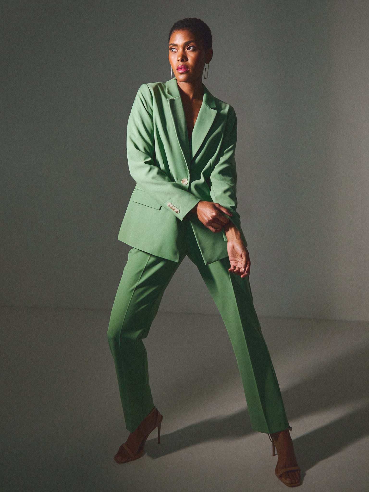 A woman dressed in a bright green suit with matching green pants, standing in a studio with a dark background. She is wearing brown high-heeled sandals and has short hair. The woman has a serious expression and is posed with her arms crossed lightly 