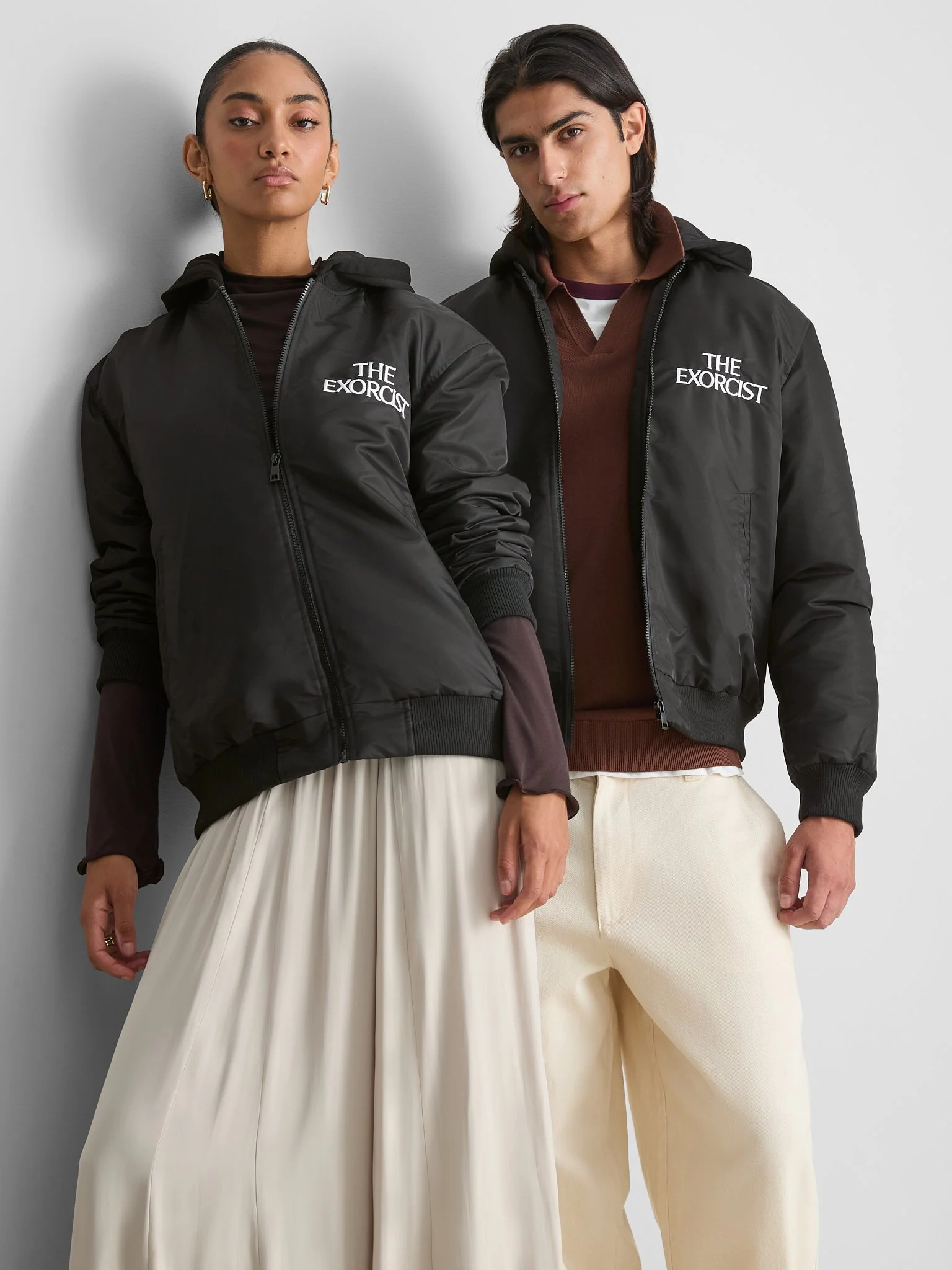 Two young adults standing against a white wall, wearing black jackets with 'The Exorcist' logo, one woman with a pleated cream skirt and a man with cream pants.