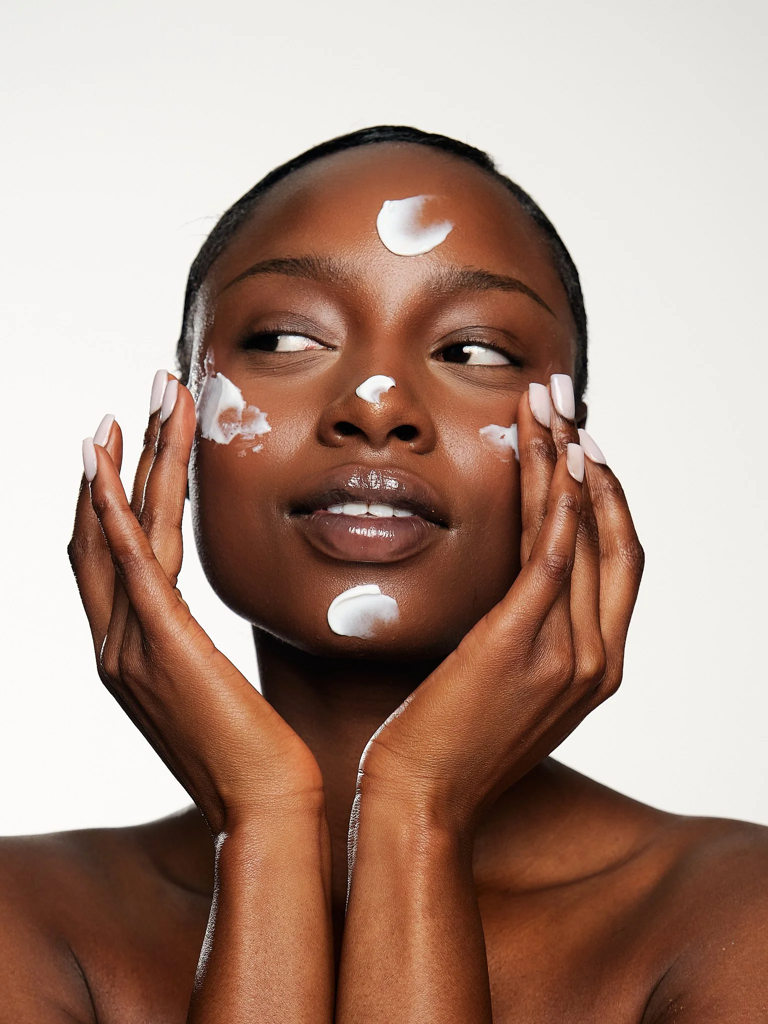 Close-up of a Black woman with skin care cream on her face and hands, against a white background.