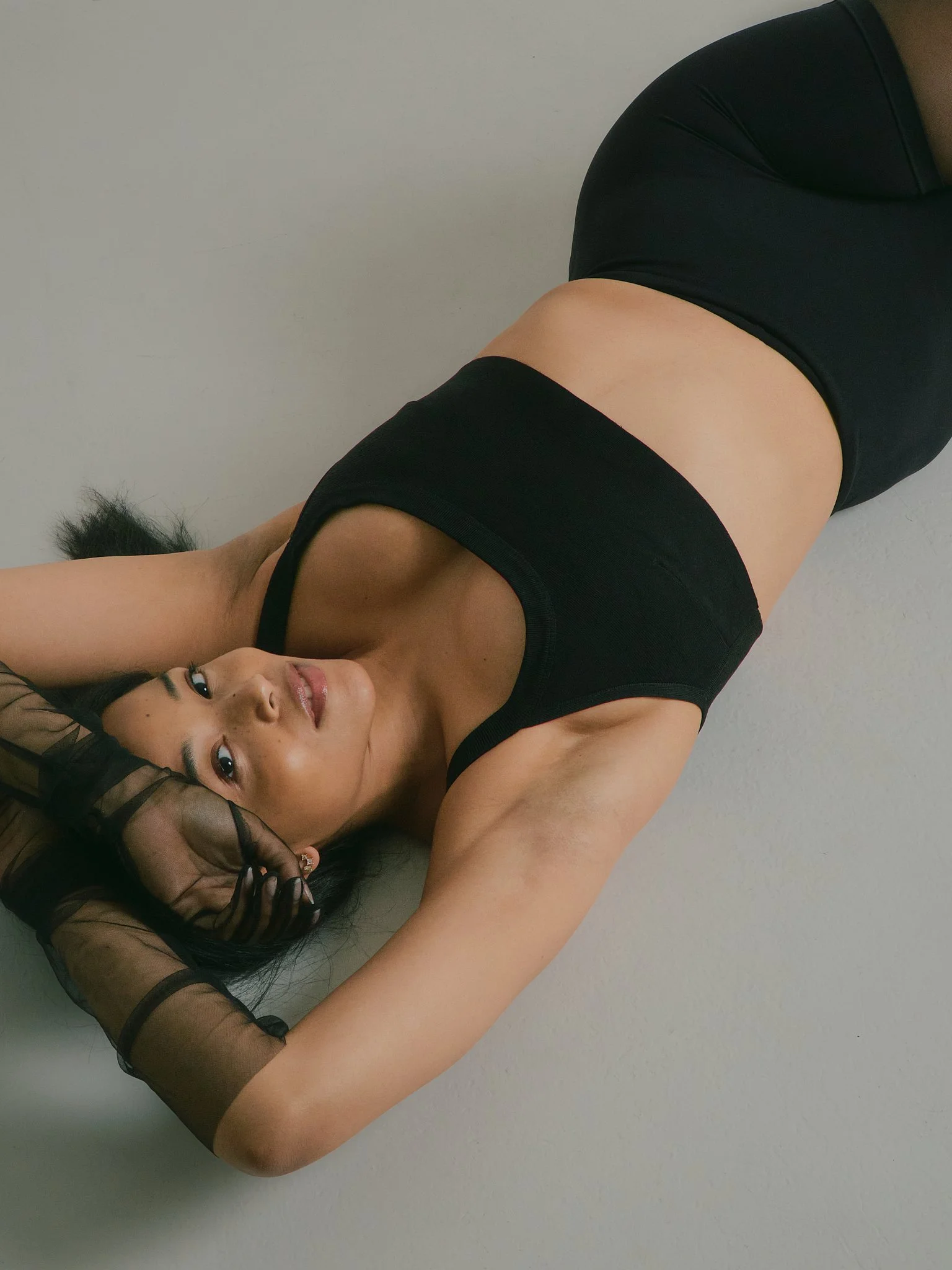 A young woman with long dark hair, wearing a black sleeveless top and gray shorts, lying on a light gray floor, looking at the camera.