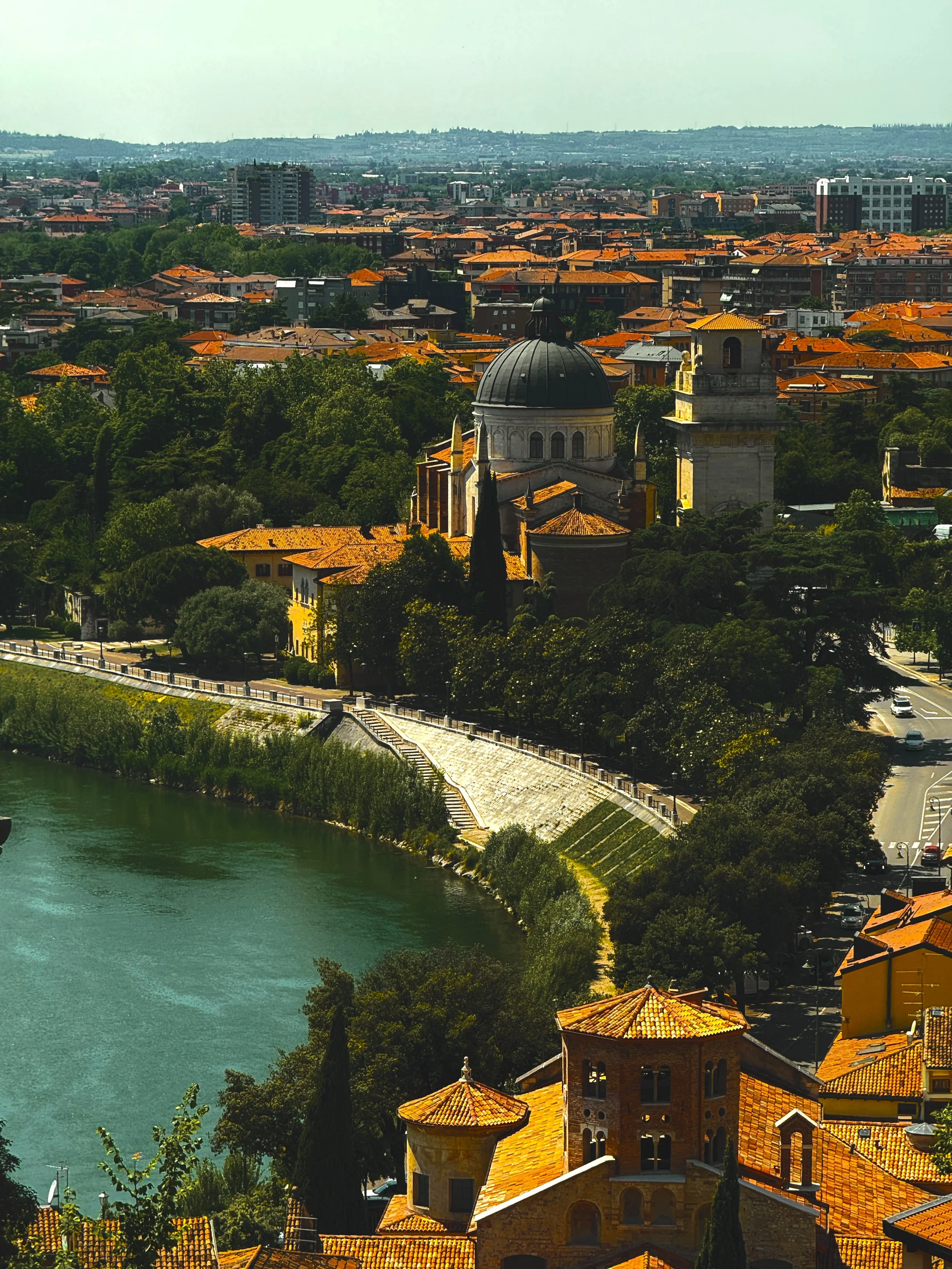 A scenic view of a European city with a river in the foreground, terracotta-tiled rooftops, historic buildings, green trees, and hills in the background.