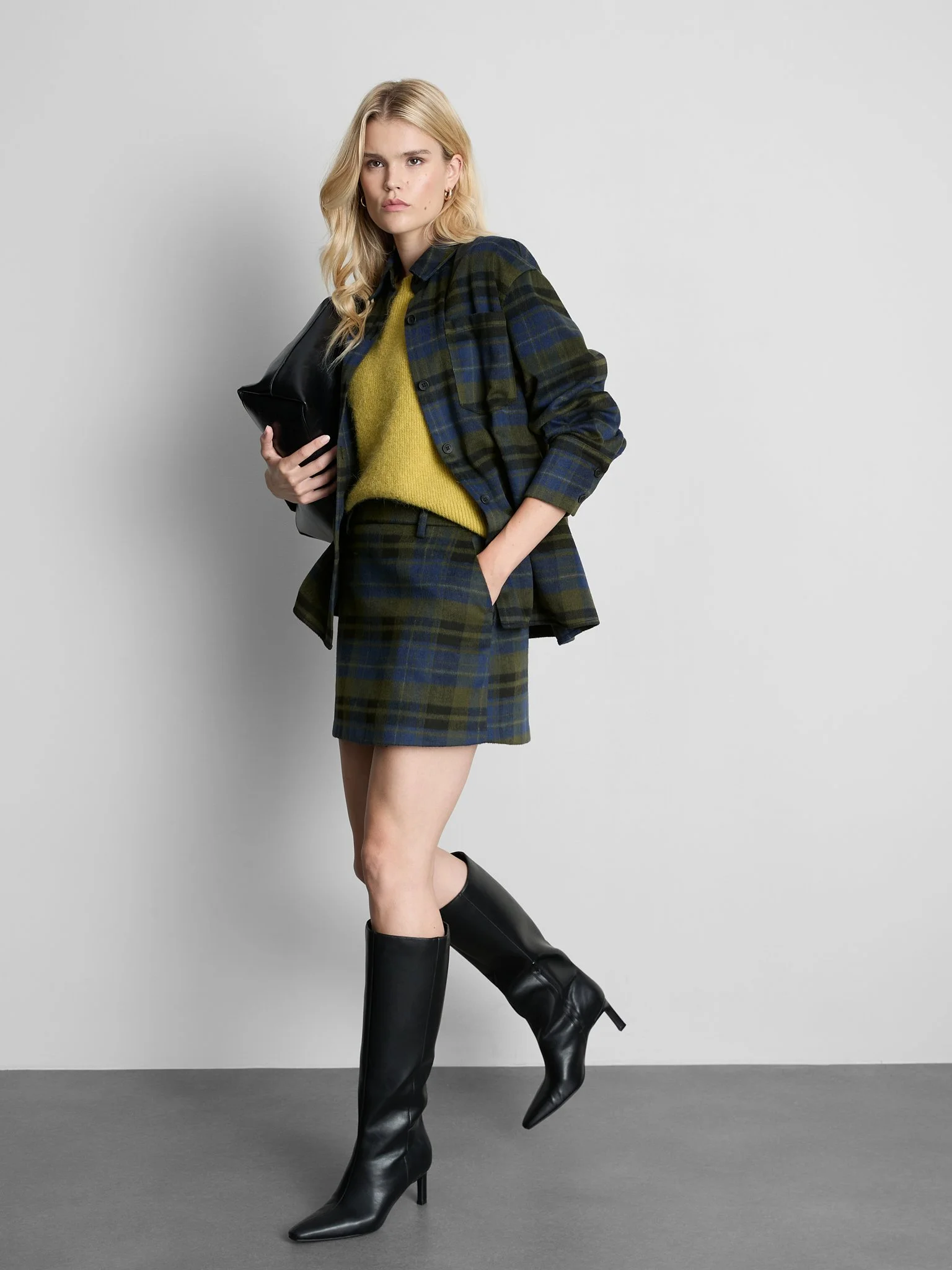 A woman standing against a plain gray wall, wearing a plaid blazer, a yellow sweater, a matching plaid skirt, and black knee-high boots.