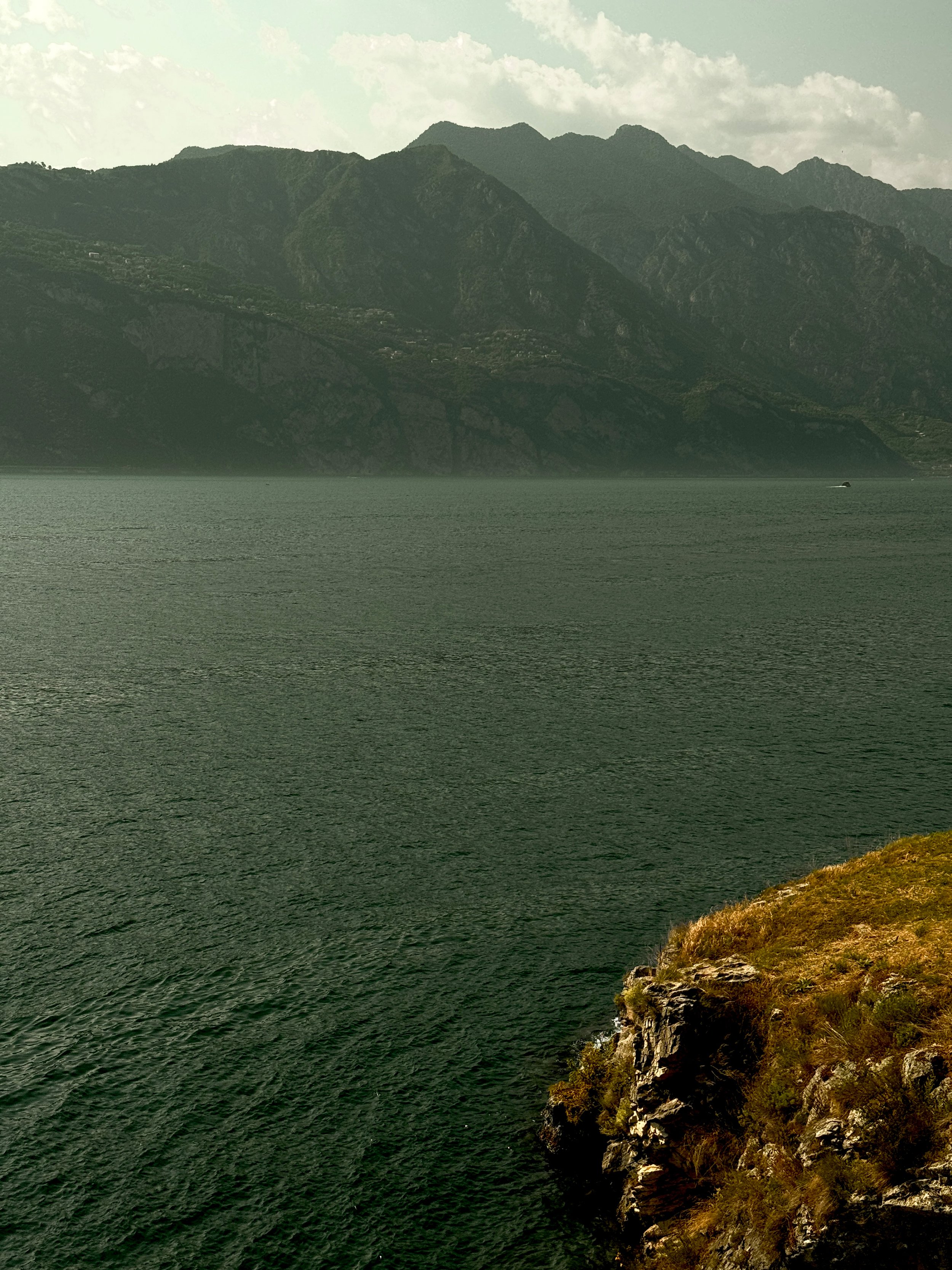 A landscape featuring a large body of water with a rocky, grassy shoreline in the foreground and a mountain range in the background under a cloudy sky.