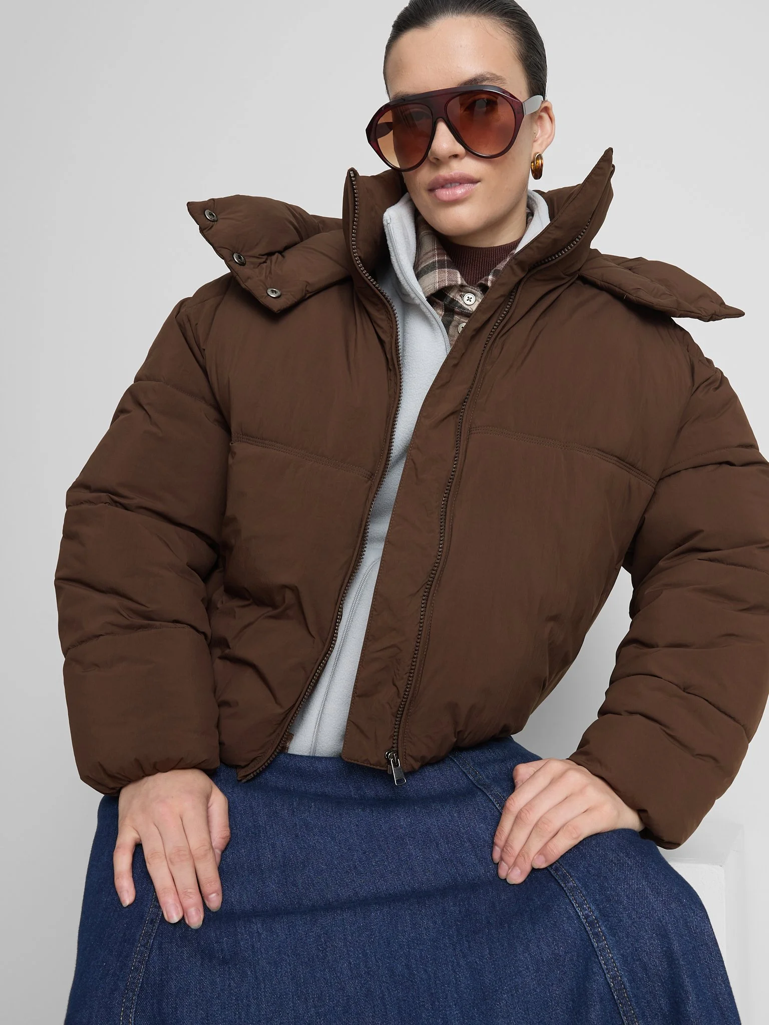 A woman wearing large sunglasses, a brown puffer jacket, and blue jeans, posing against a plain background.