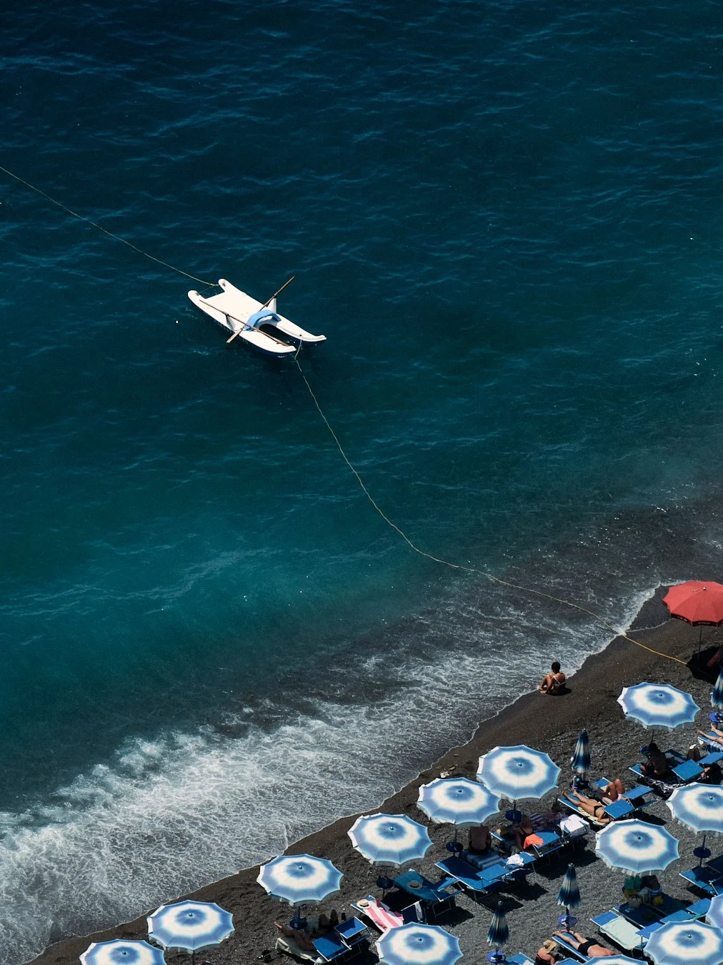 A beach scene with a white boat floating in the water near shore, close to several blue and white umbrellas and people relaxing on a rocky beach.