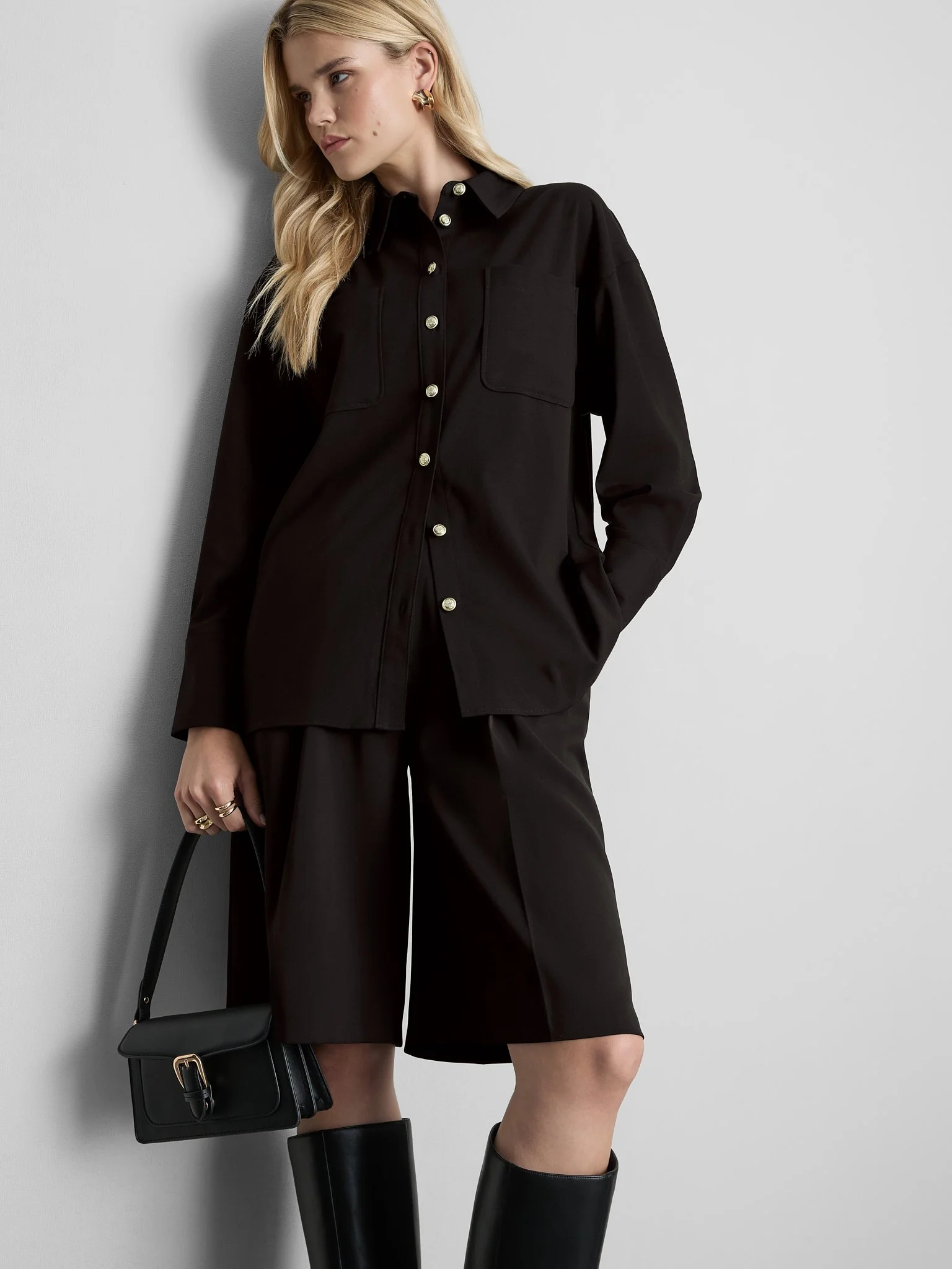 A woman with blond hair wearing a black button-up coat, black skirt, and black knee-high boots, holding a small black handbag with a gold buckle, standing against a plain light gray background.