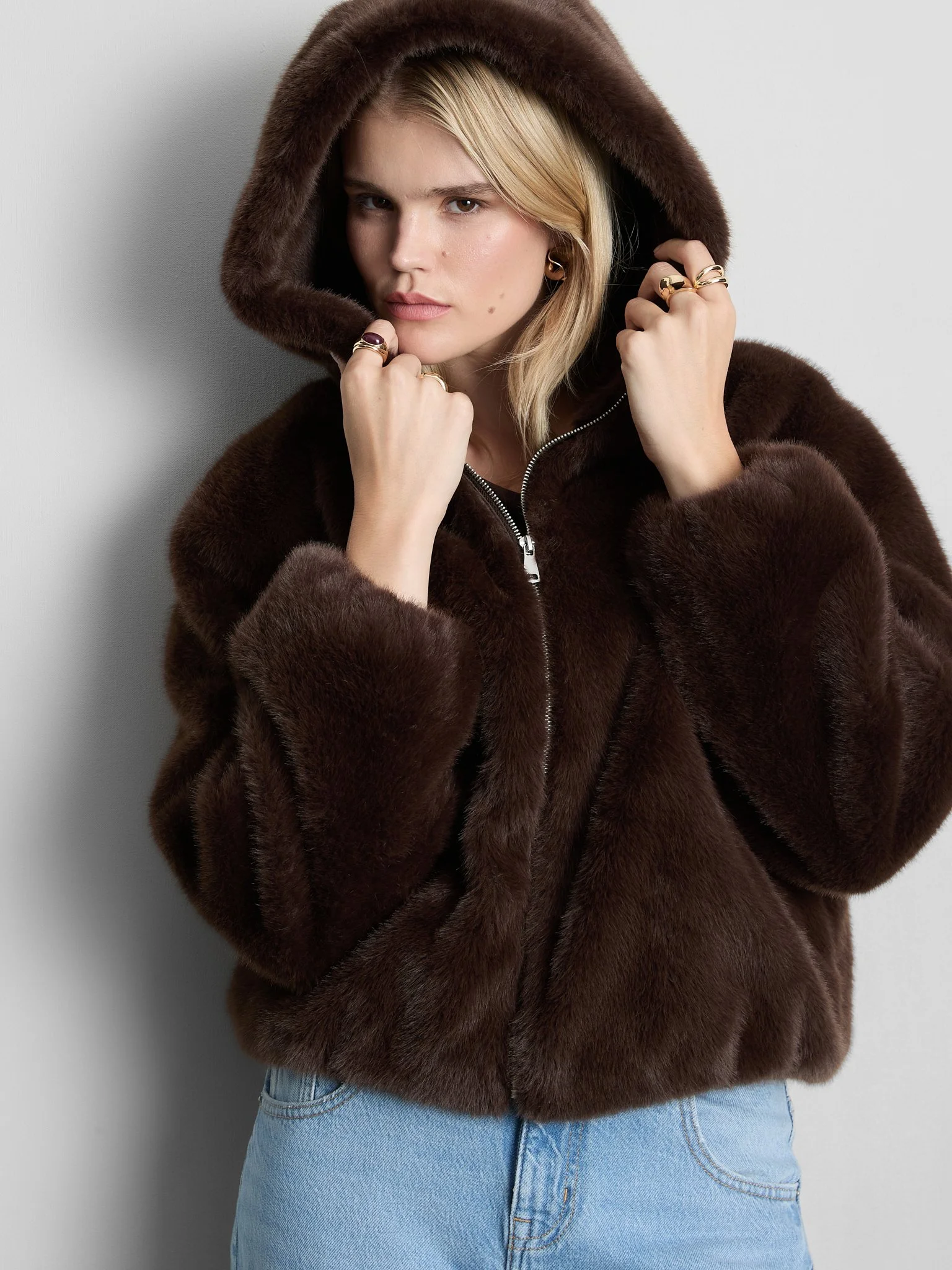 A person wearing a brown faux fur hooded coat and light blue jeans, standing against a plain light background.