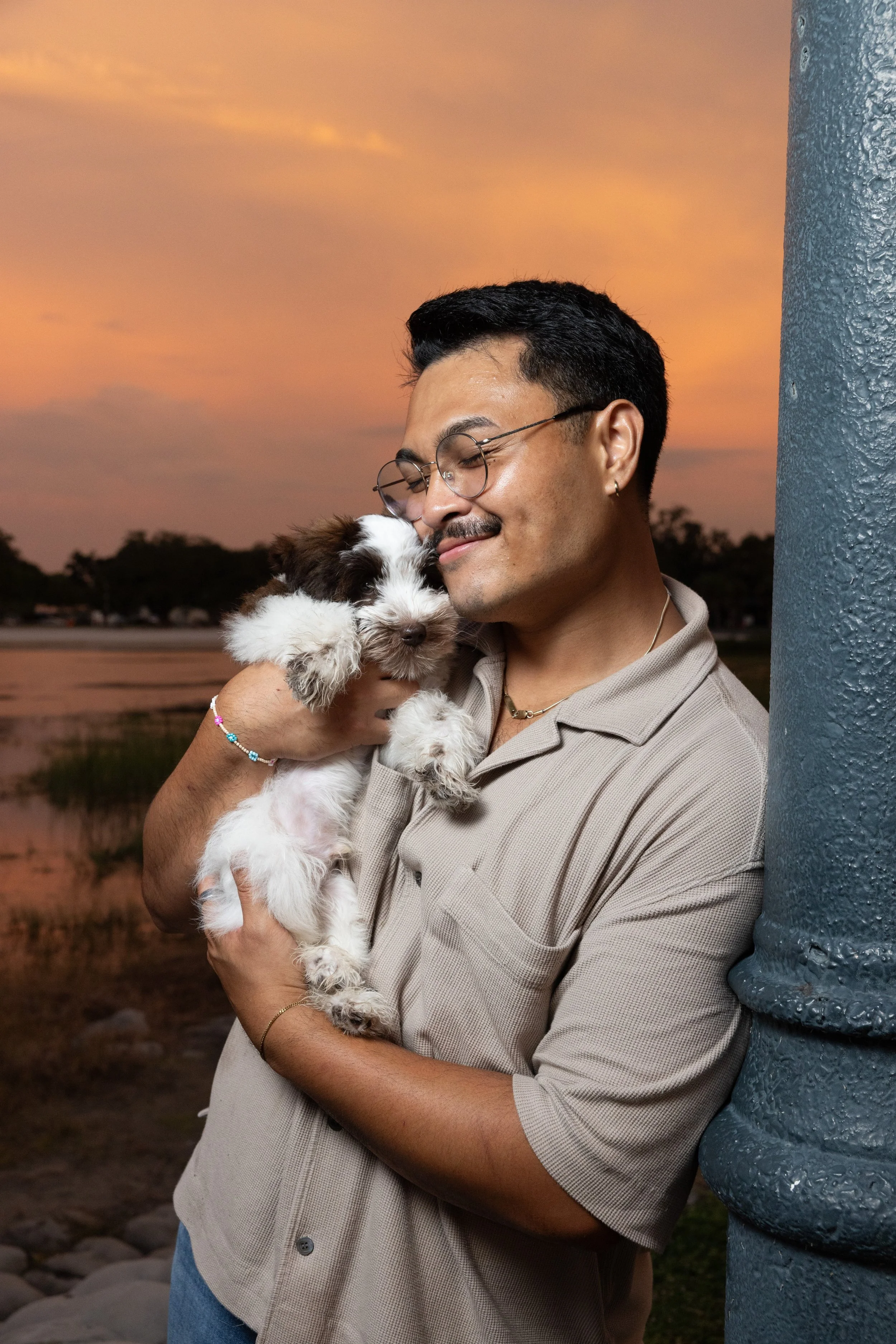 A man with glasses and a mustache holding a small black and white puppy close to his face outdoors at sunset.