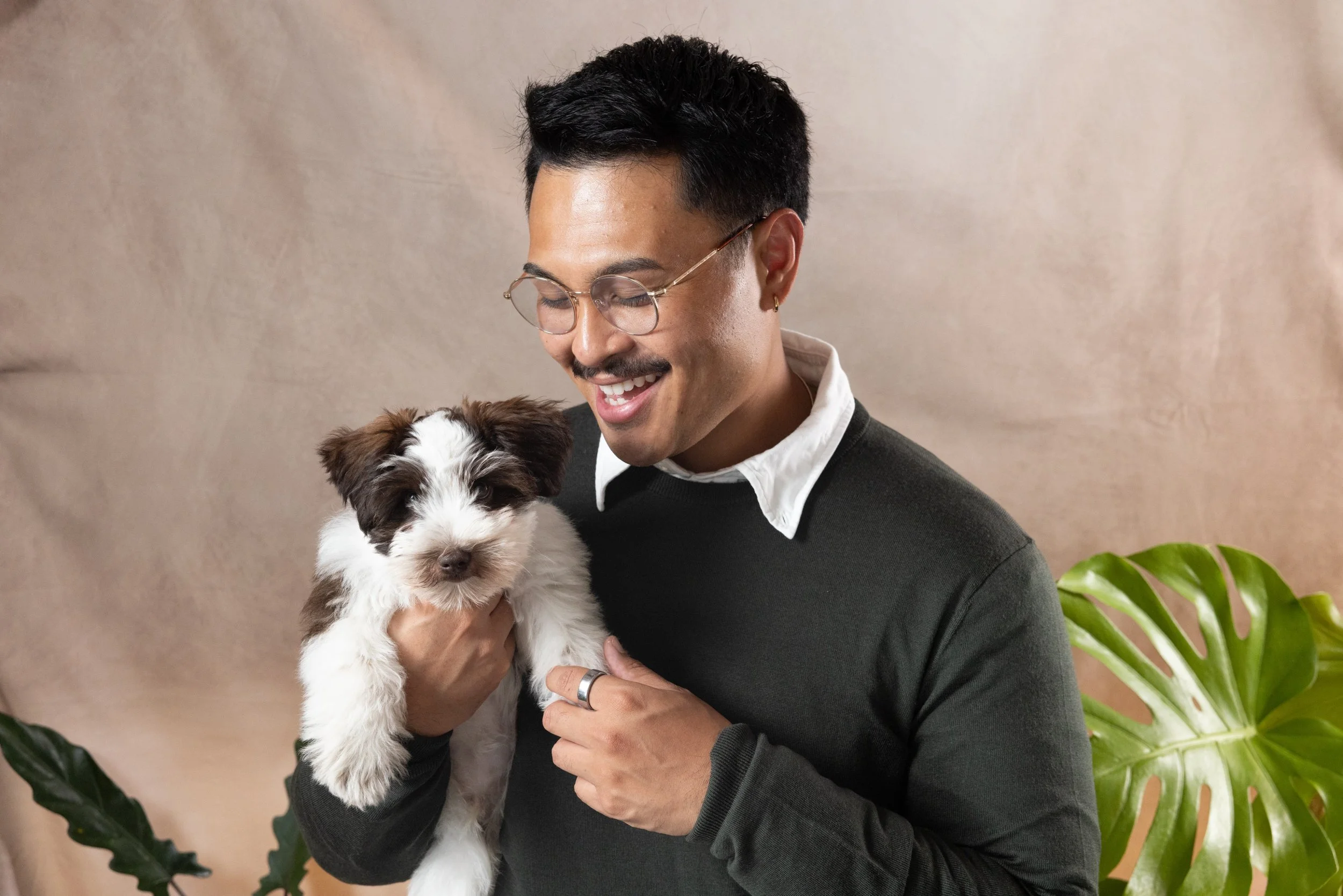 A man with dark hair, glasses, and a mustache is smiling as he holds a small, fluffy black and white puppy. There are large green houseplants behind them and a neutral-colored background.