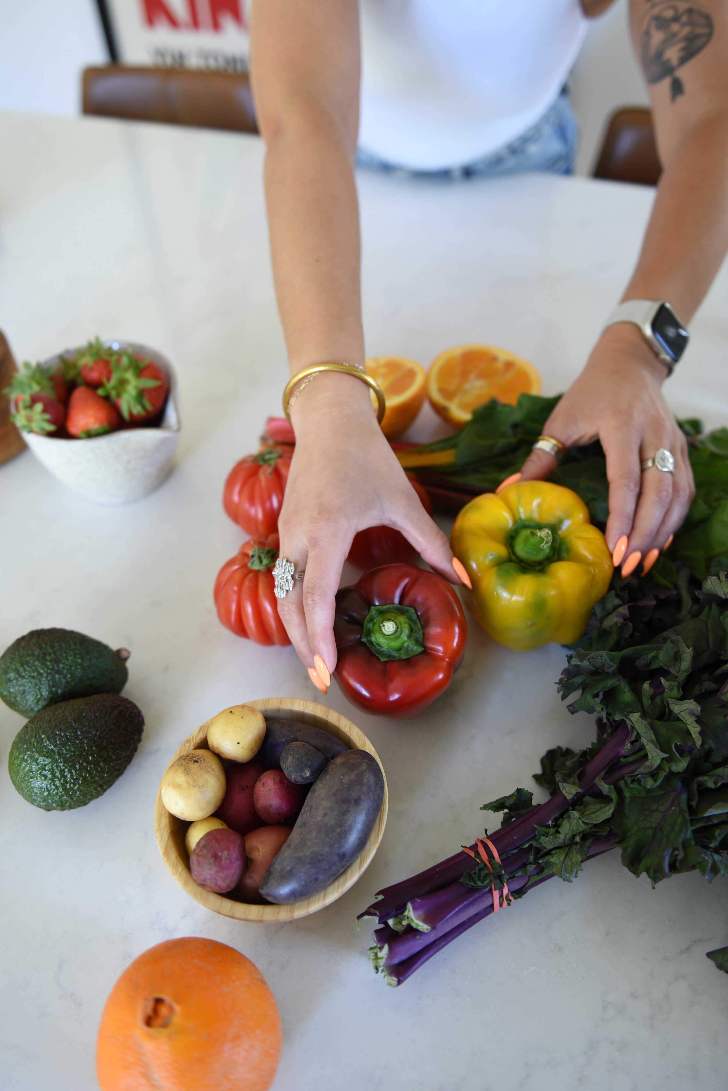 Kitchen countertop filled with fruits and vegetables. Liv reaching for a pepper.