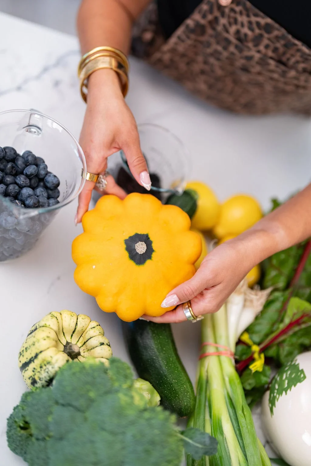 Person handling yellow and green squash and placing it with other fresh vegetables including blueberries, cucumbers, and squash on a white surface.
