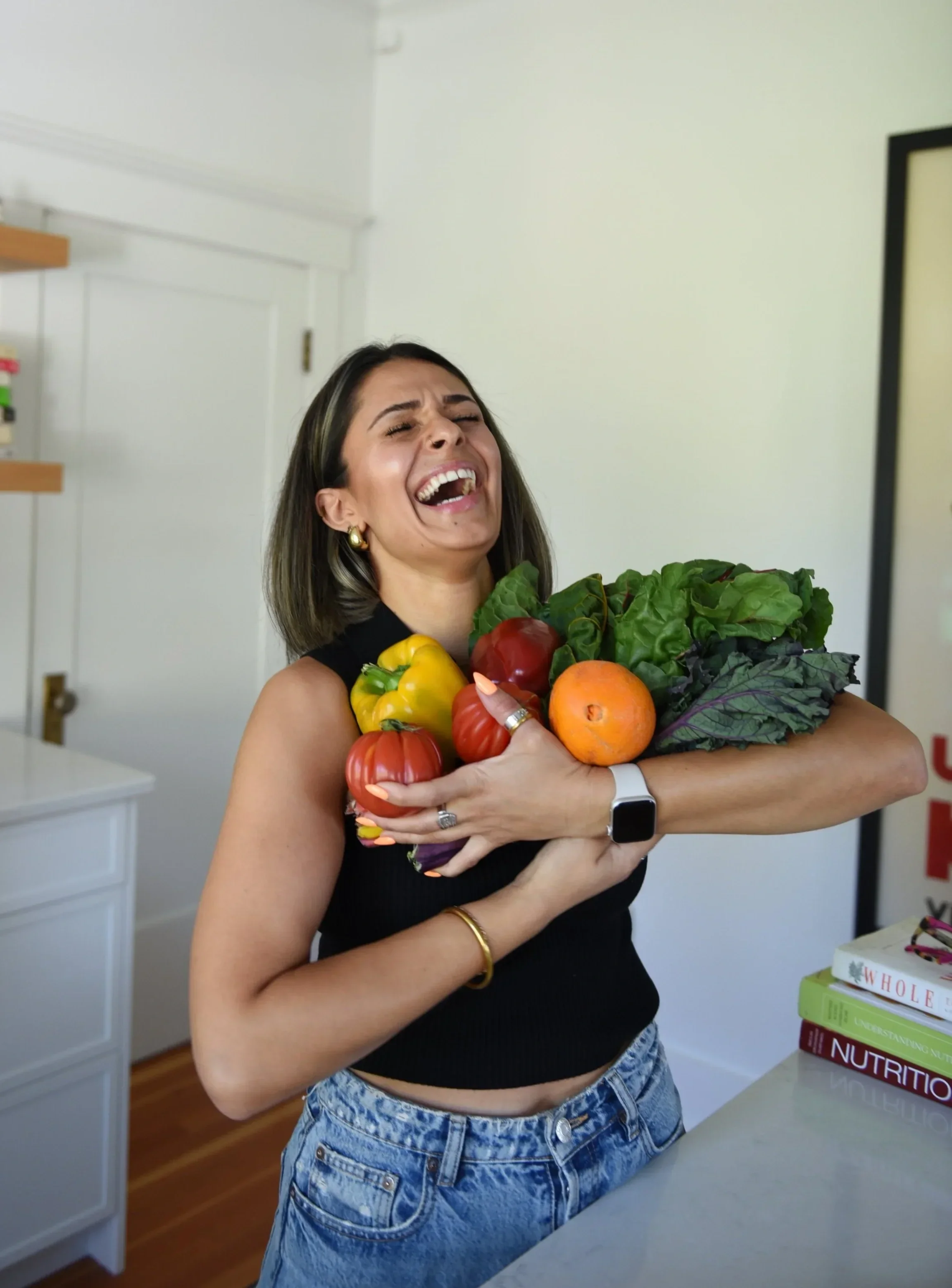 A woman (Liv) with shoulder-length hair, gold earrings, and a black sleeveless top is smiling and hugging a large bunch of colourful fresh fruits and vegetables in a kitchen.