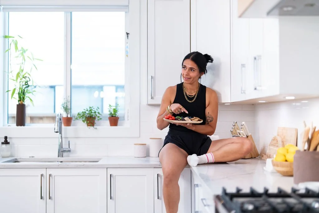 A woman sitting on a kitchen counter holding a plate of food, with sunlight streaming through a window behind her.