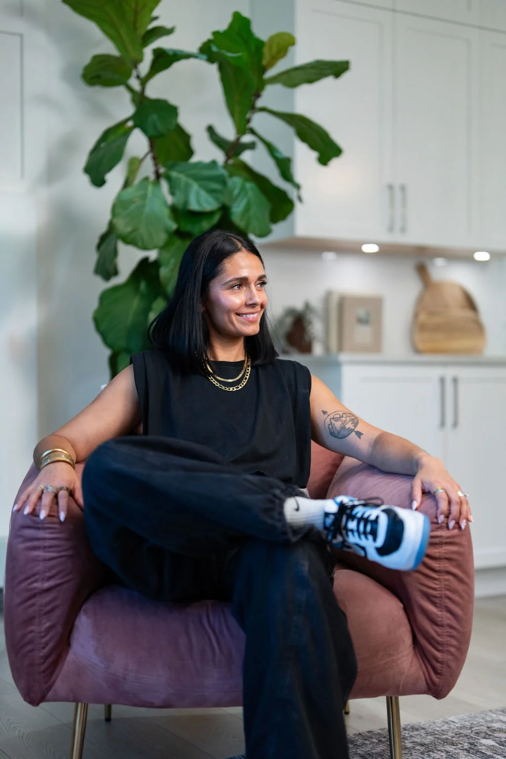 A woman with black hair and tattoos relaxing on a pink armchair in a modern, bright kitchen with green plants and white cabinets.