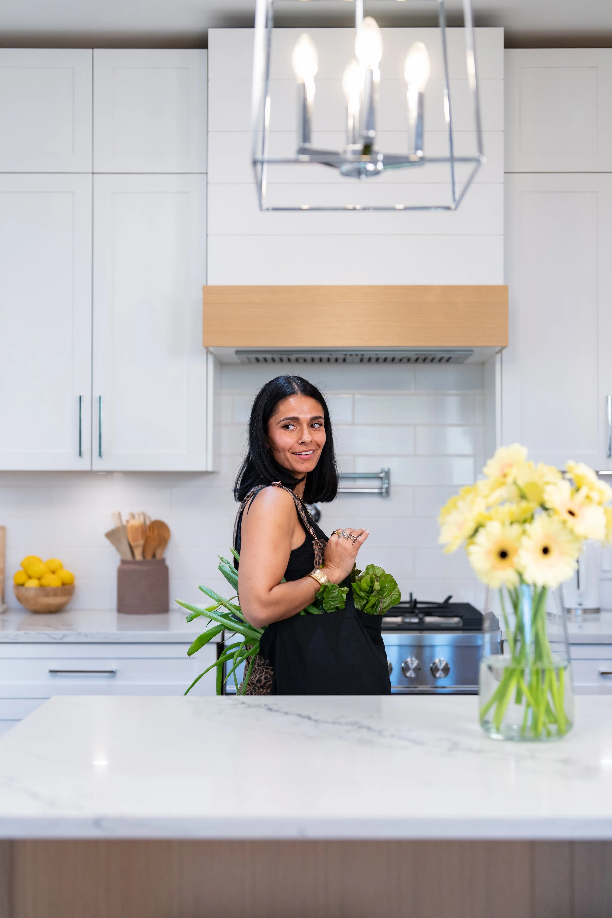 A woman with black hair and a black apron standing in a modern kitchen, holding fresh green vegetables, with a vase of yellow flowers on the white marble countertop in the foreground.