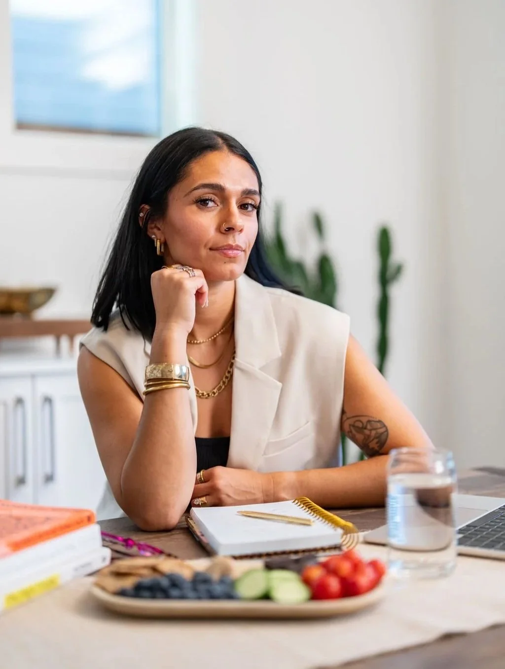 Olivia (Liv) Rahal at her desk with her agenda, water, and fresh fruits and vegetables. She has one arm crossed and the other is proped up on the table with her head resting on her hand