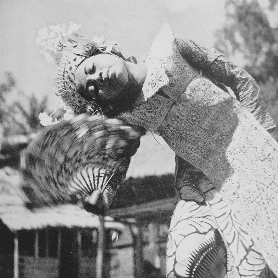A classic photo of a woman in traditional Balinese clothing while holding a fan and dancing outdoors.