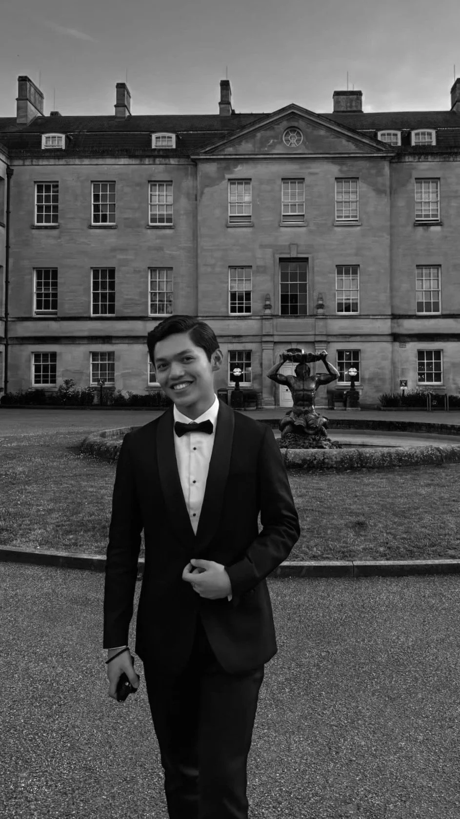A young man in a tuxedo standing in front of a historic building with a fountain, smiling for the camera. Bumi Adventures Founder. Julio Iskandar.