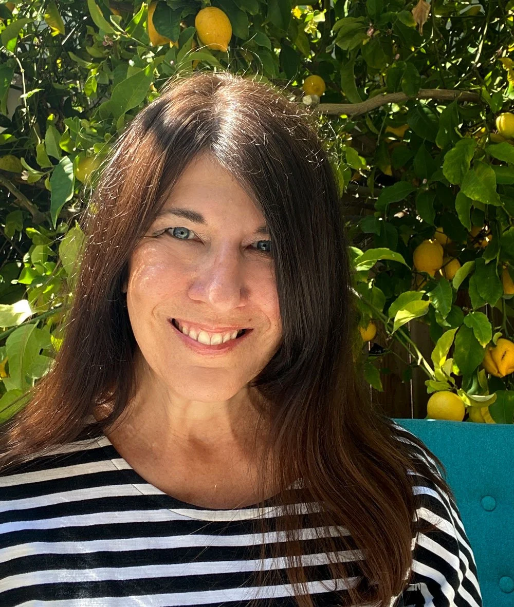 A portrait of Lisa Bellew smiling outdoors in front of a lemon tree with yellow lemons and green leaves, wearing a black and white striped shirt.