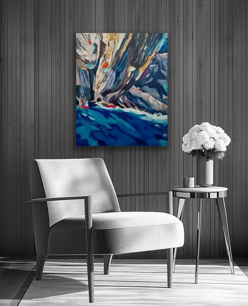 A modern interior with a black wooden wall, a framed abstract painting by Lisa Bellew with shades of blue and beige, a white upholstered armchair, a small round side table with a white vase of white flowers, and a candle.