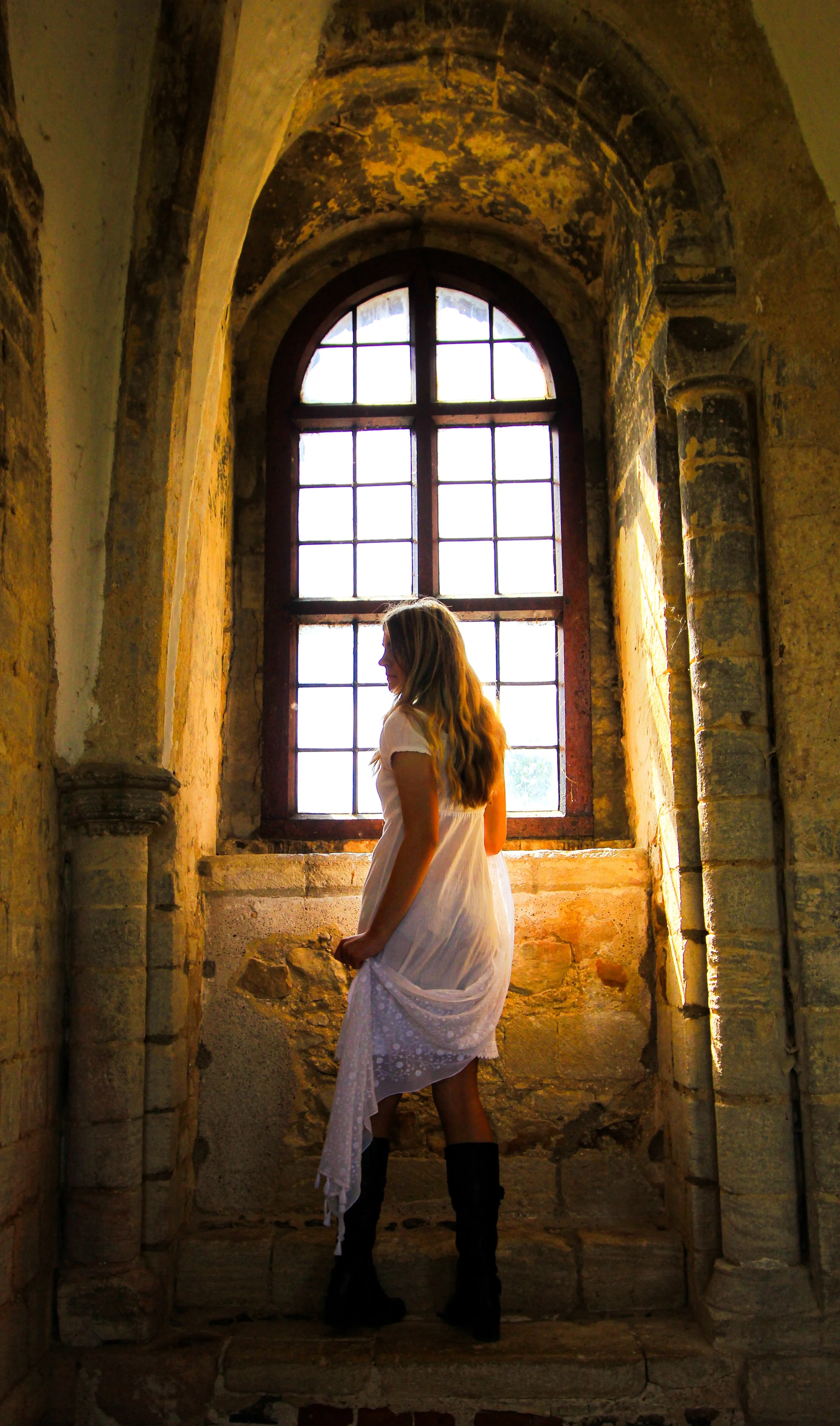 A woman with long blonde hair wearing a white dress and black knee-high boots standing inside an old stone building with a large arched window behind her.