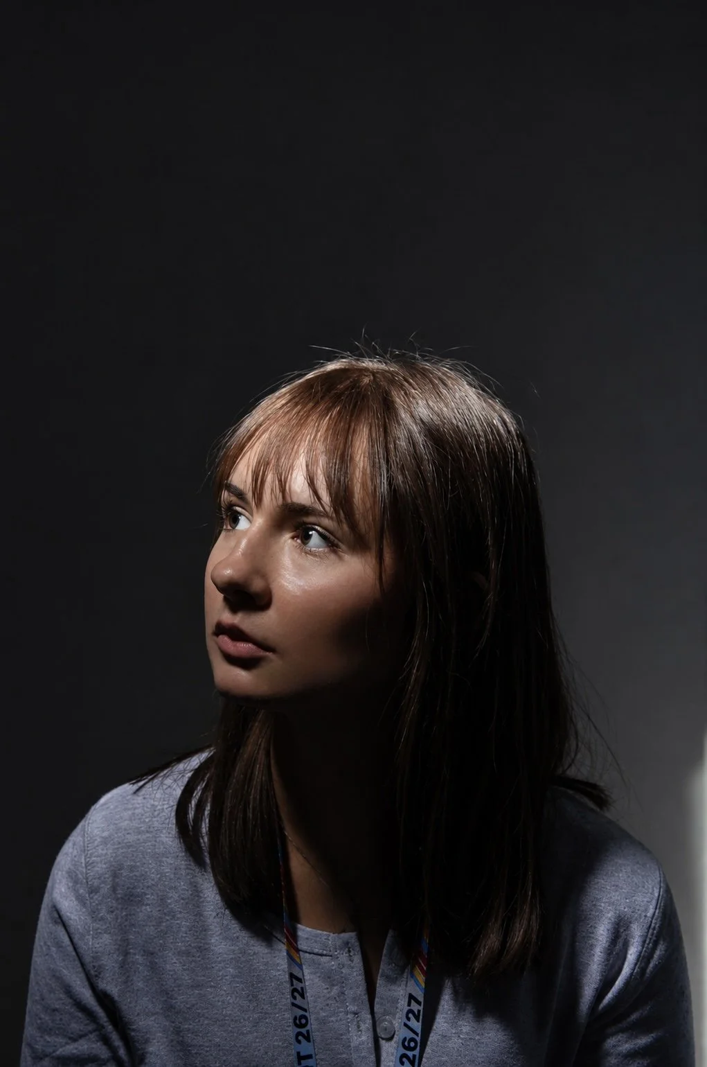 A young woman with brown hair and fair skin looking up and to her left against a dark background, wearing a gray shirt and a lanyard with dates 26/12 and 27.