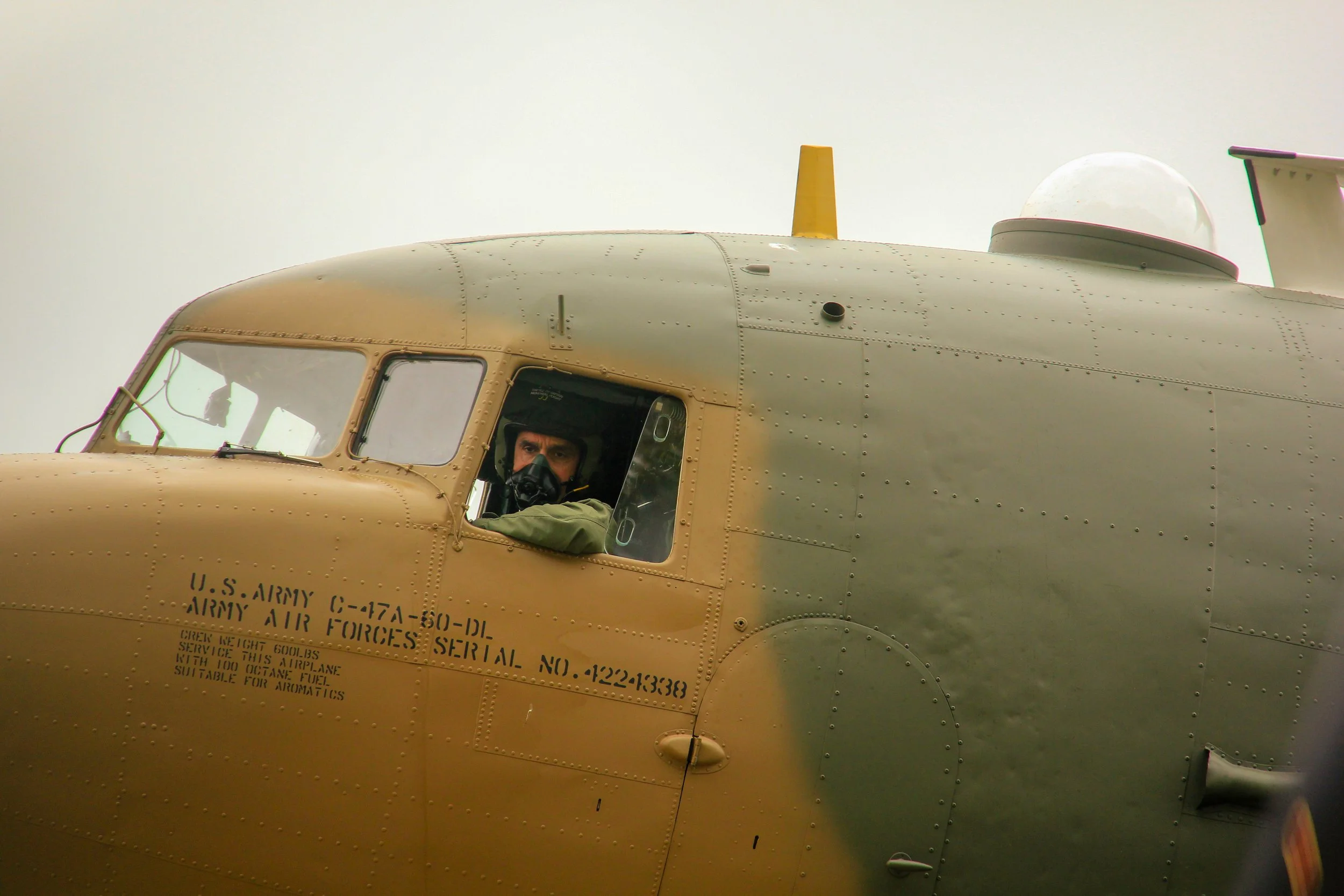 Close-up of a U.S. Army helicopter cockpit with a pilot inside, wearing a helmet and oxygen mask, partially visible through the window. The helicopter's exterior is tan with visible rivets and markings.