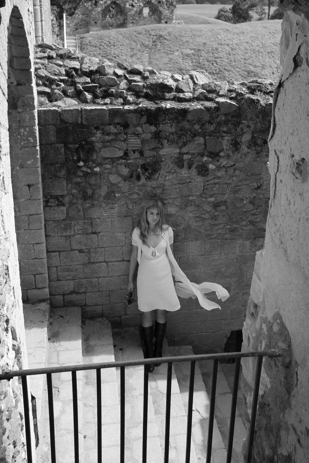A young woman in a white dress and tall black boots standing on stone steps between two stone walls, holding a small object in her right hand, with long hair and a neutral expression.