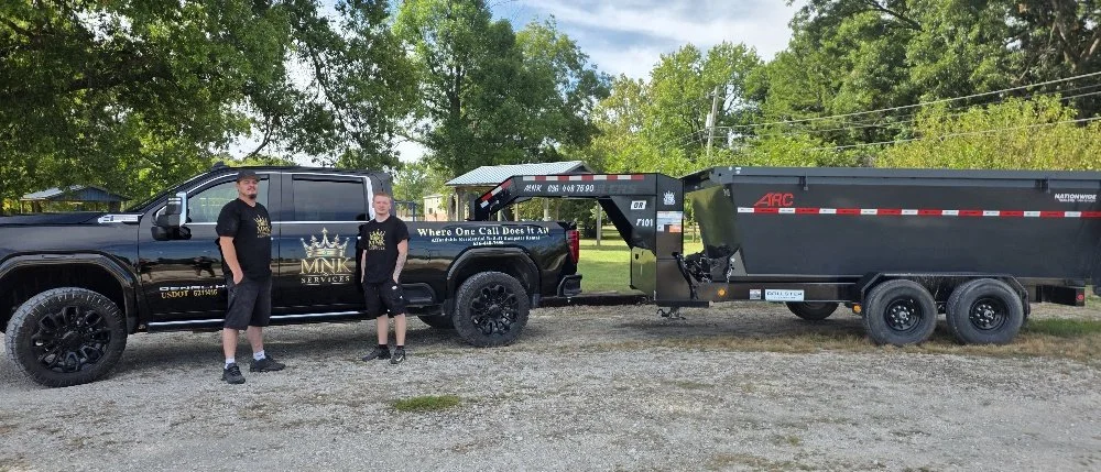 Two men standing near a black pickup truck with a logo for MNK Services parked on a gravel lot. The truck is towing a large black trailer.