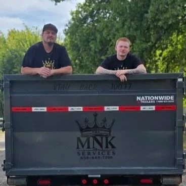 Two men standing behind a black trailer with company logos and contact information, outdoors with trees in the background.