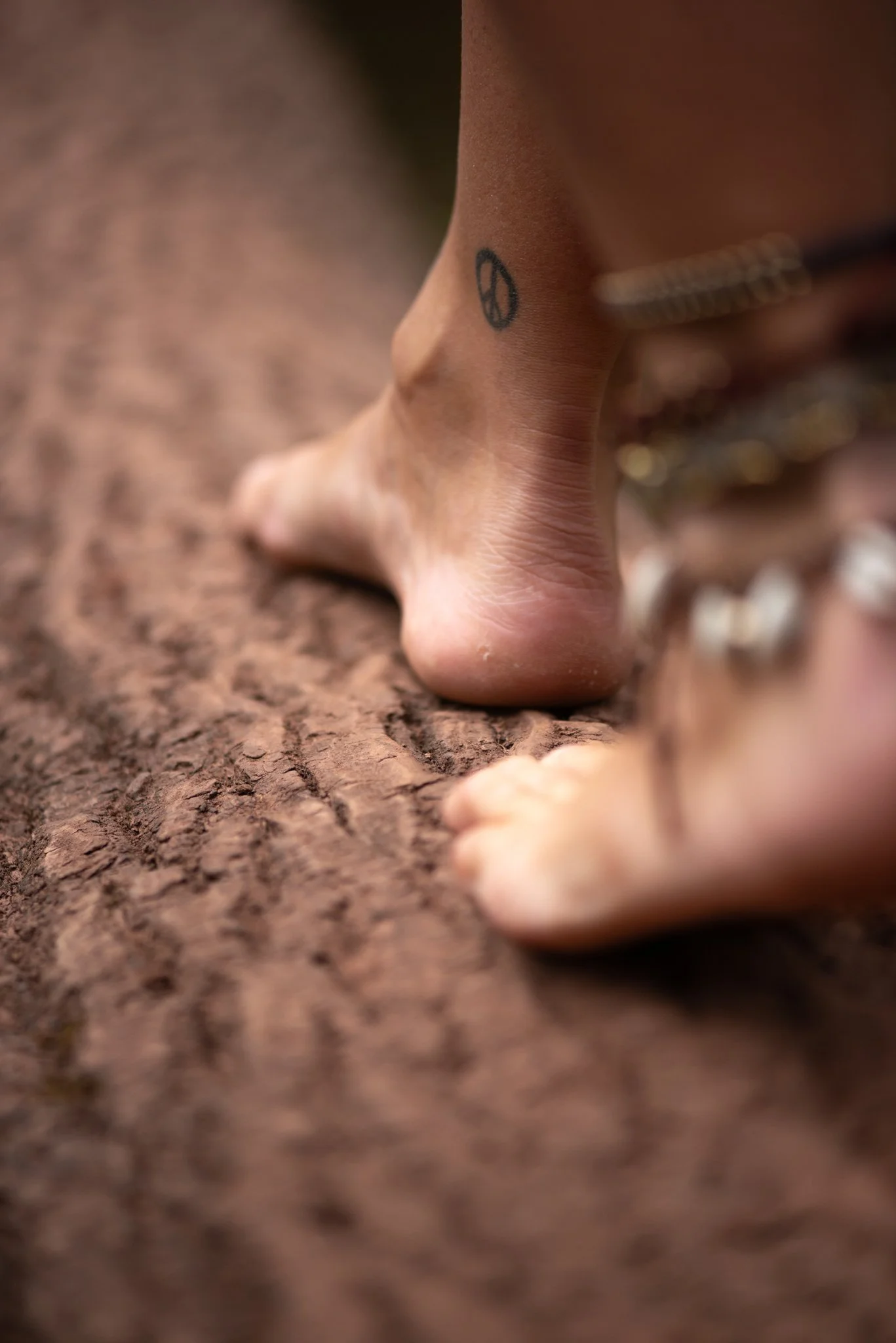 Close-up of a person's foot with a peace sign tattoo on the ankle, standing on textured ground with blurred pebbles in the background.