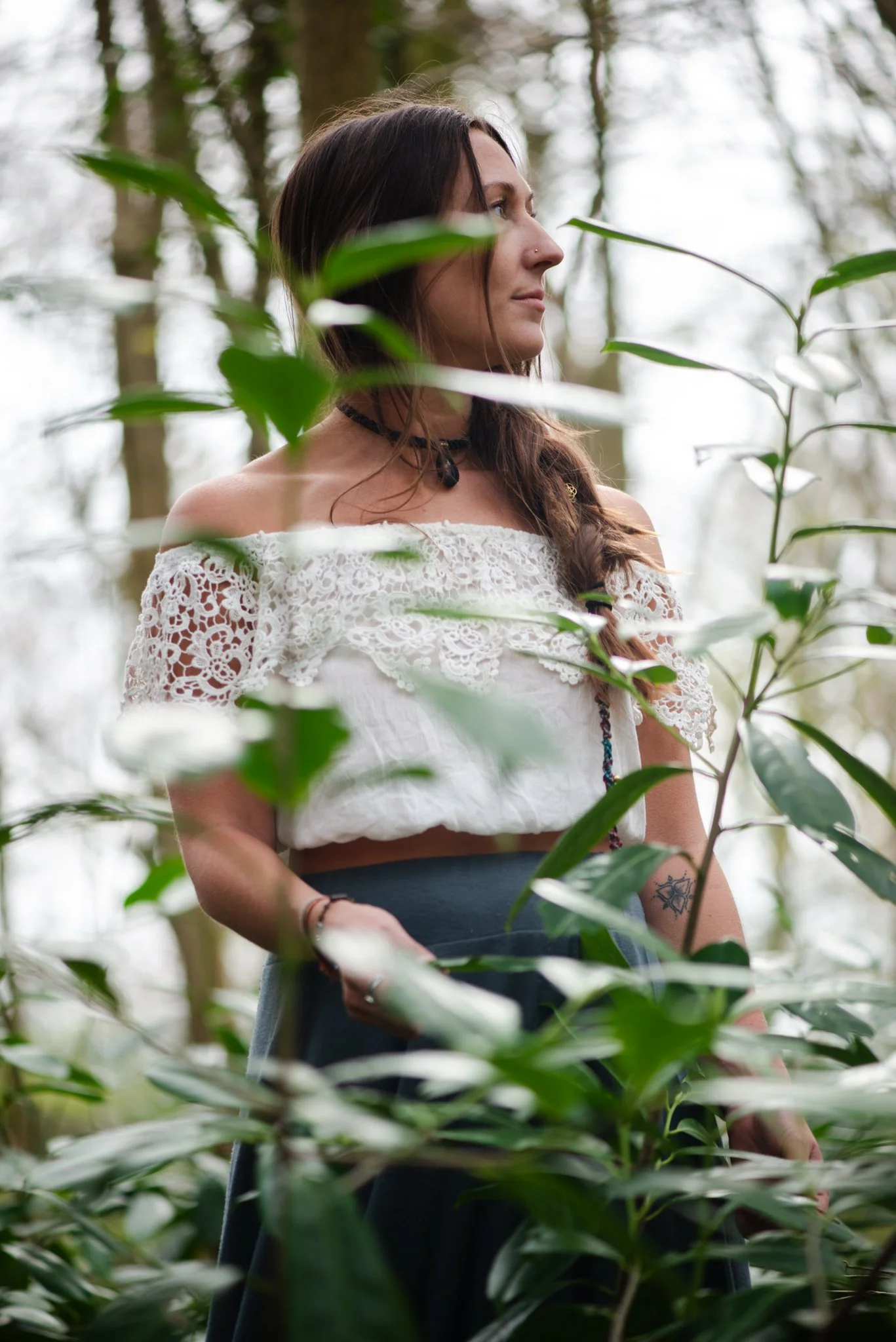 A woman with long brown hair and a nose piercing wearing a white lace off-shoulder top, standing among green plants in a forest or garden, with blurred trees in the background.