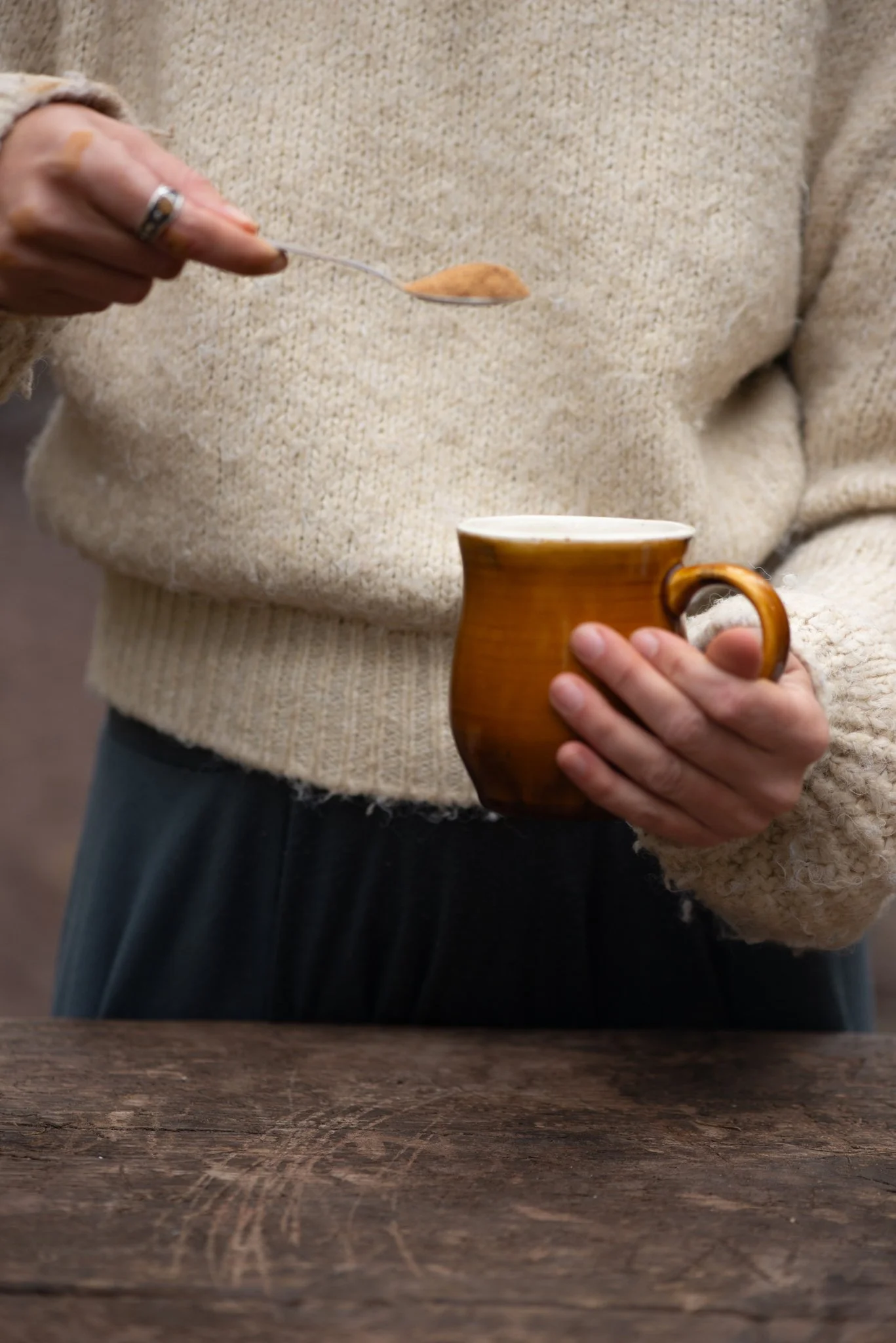 Person wearing a beige sweater and dark pants holding a rustic brown mug in one hand and a spoonful of powder in the other, with a wooden table in the foreground.