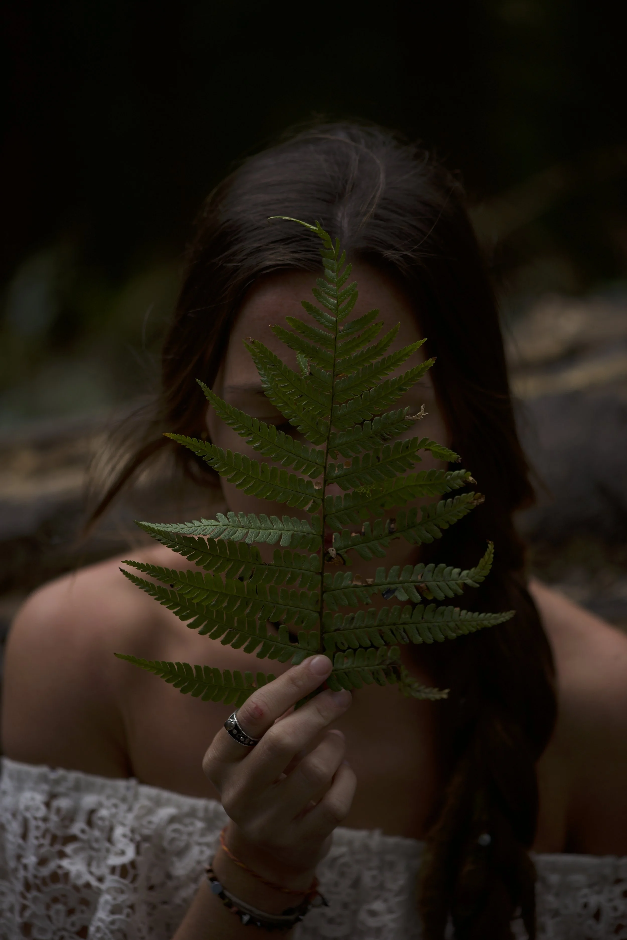 A woman holding a green fern in front of her face, with dark hair, wearing a white lace top, and jewelry on her fingers and wrist.