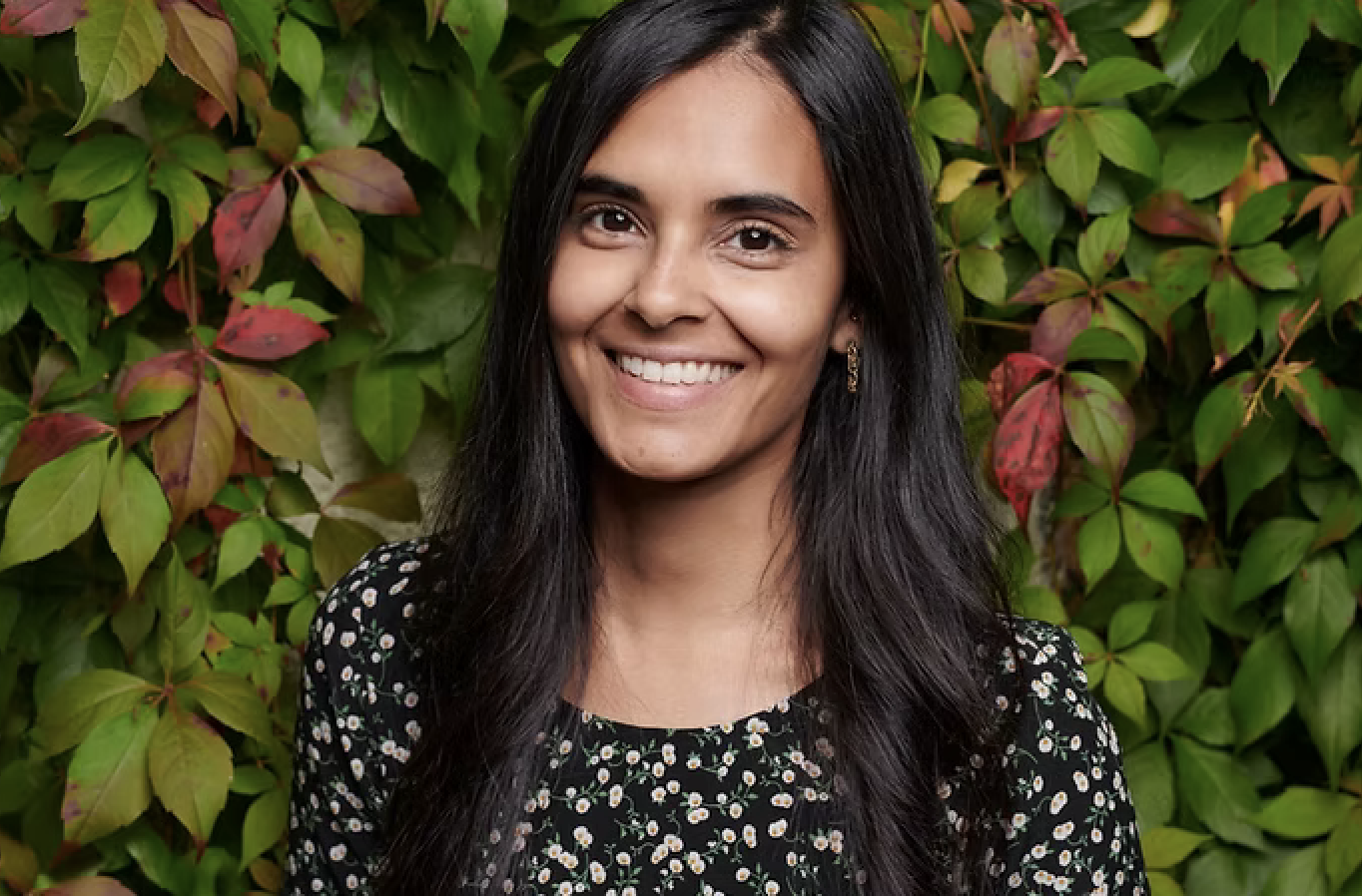 A woman smiling outdoors in front of green and reddish leaves.