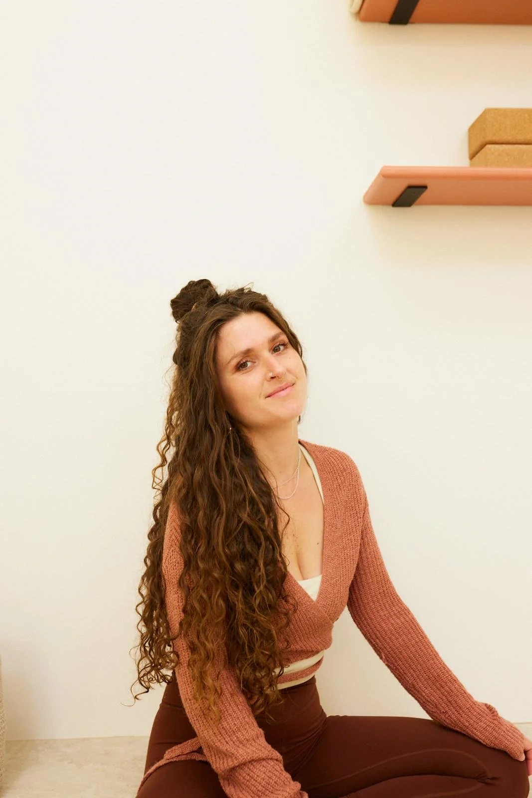 Young woman with long, curly brown hair sitting on the floor against a plain wall, wearing a pinkish-brown sweater and brown pants, with shelves and boxes visible behind her.