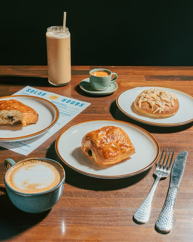 A breakfast scene with two plates of pastry, a cup of coffee, a latte, and a chocolate drink on a wooden table.