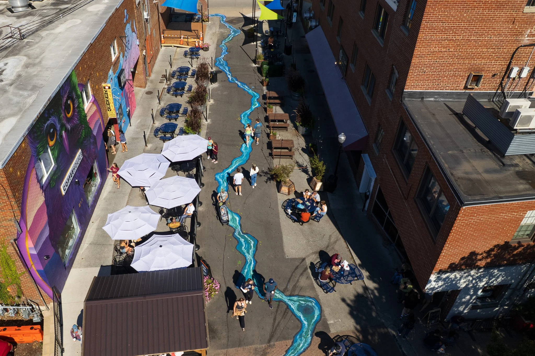 Downtown outdoor cafe with blue tables and umbrellas, a painted mural on a building wall, a painted water stream on the ground, and people walking and sitting.