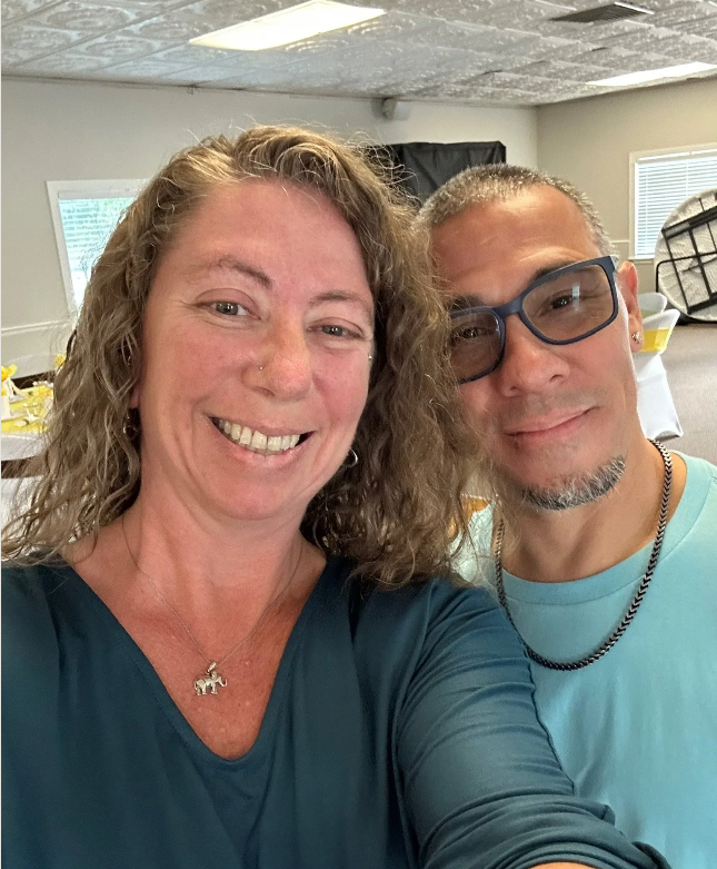 A smiling woman with curly hair wearing a navy blue top and a necklace with a dog pendant, taking a selfie with a man wearing glasses, a light blue shirt, and a chain, in a room with tables, chairs, and a window.