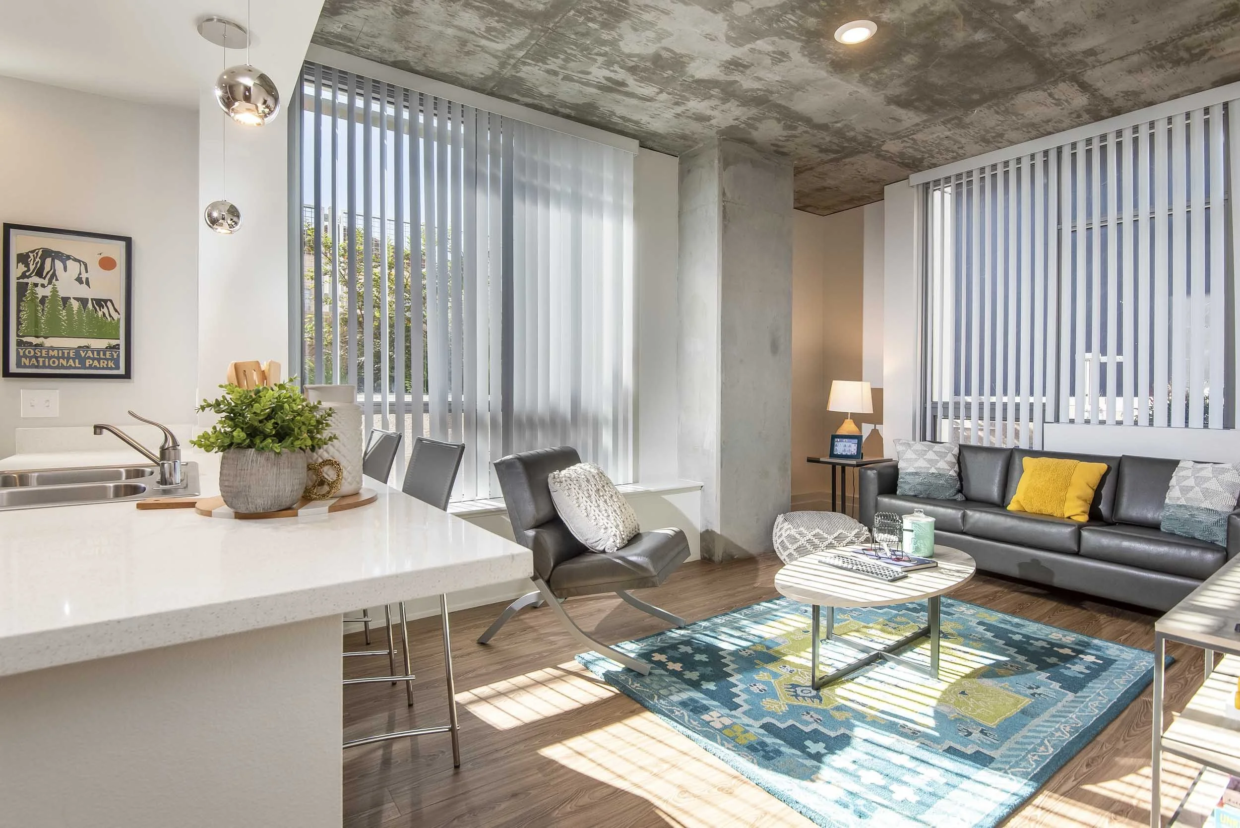 Living room with large windows covered by vertical blinds, a black leather sofa with yellow and patterned pillows, a floor lamp, a coffee table with green books, a modern area rug, and a portion of a kitchen island with a potted plant.