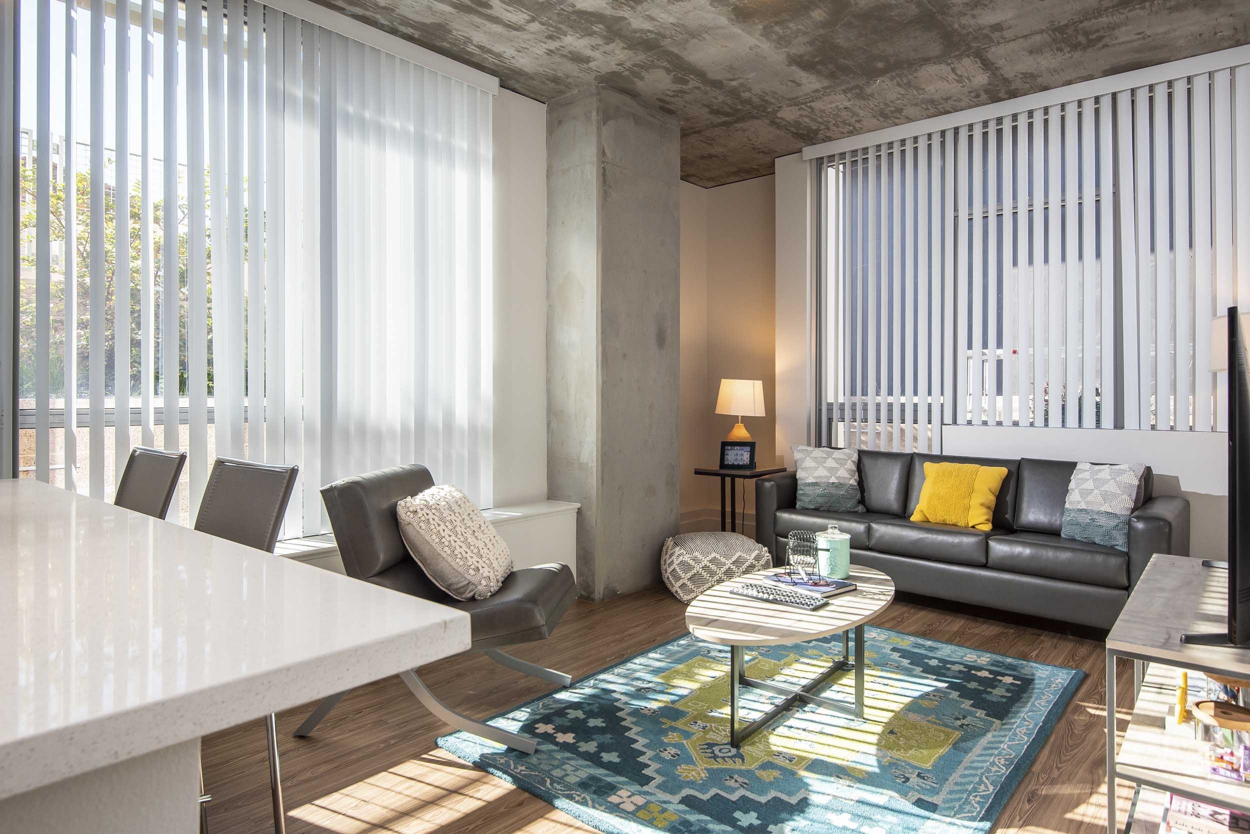 Modern living room with large windows, vertical blinds, a concrete ceiling, a gray sofa with yellow and gray cushions, a round coffee table, a floor lamp, and a colorful area rug.