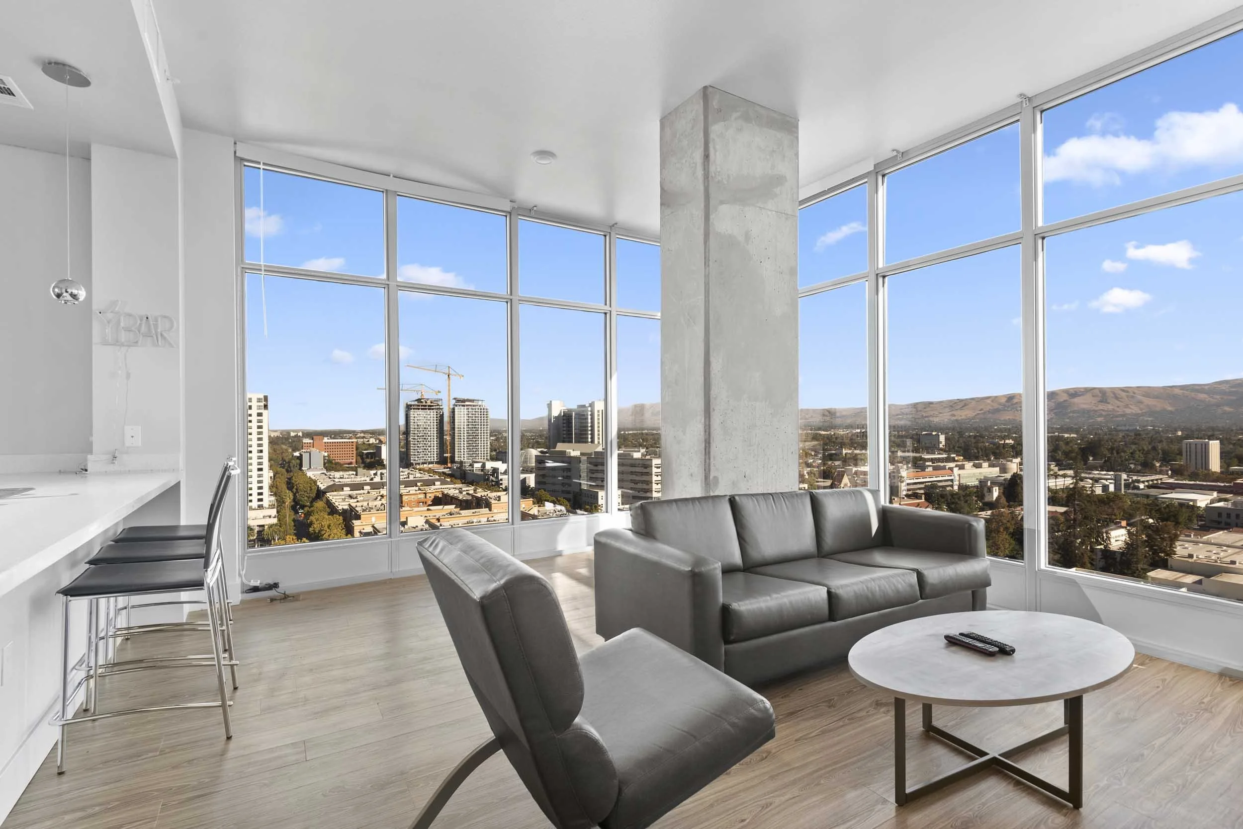 Modern living room with large floor-to-ceiling windows showing a cityscape and mountains, featuring a gray sofa, a round coffee table, and bar stools at a white counter.