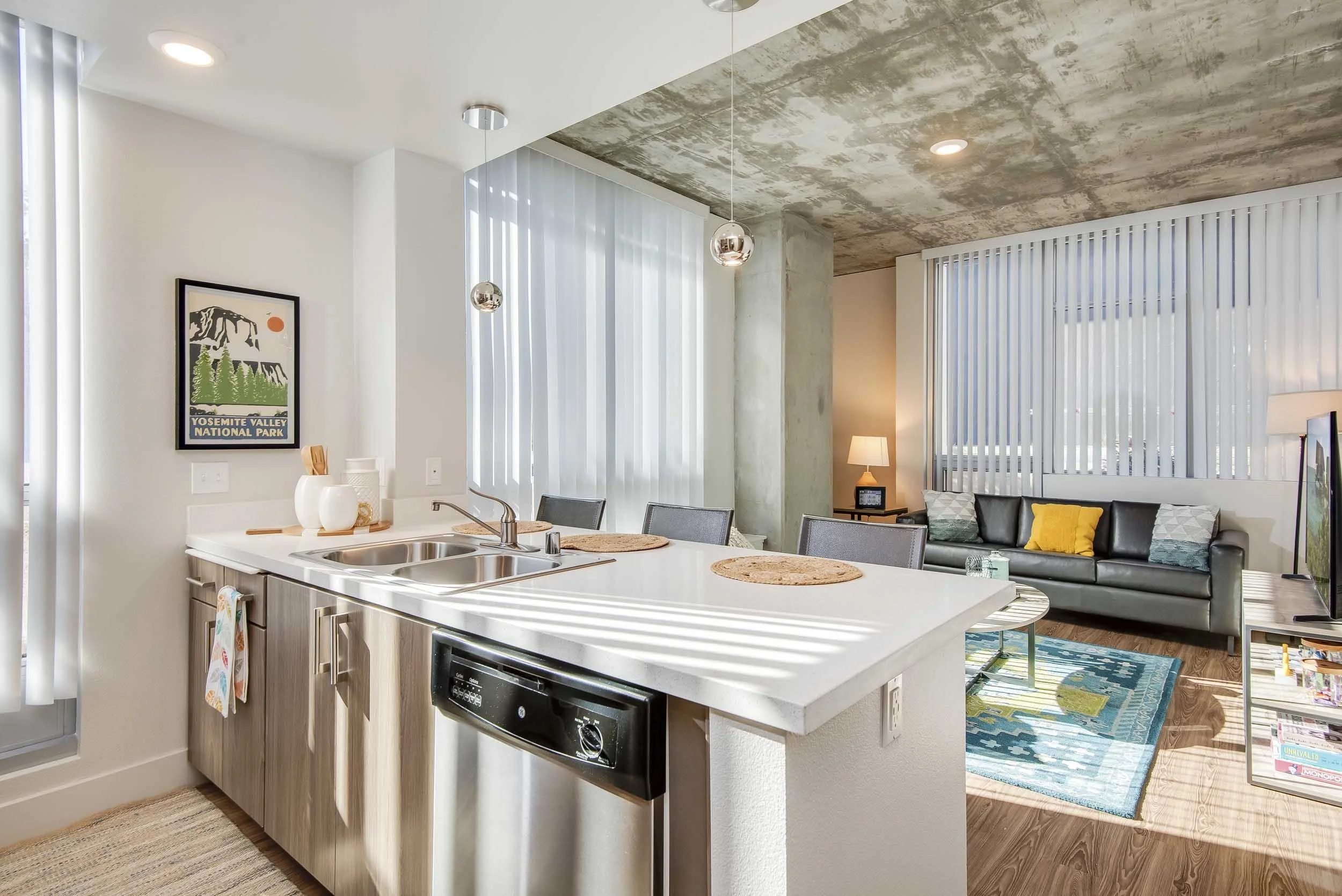 Modern open-concept living space with a kitchen and living room. The kitchen has a white island with a double sink, light wood cabinetry, and woven placemats. A framed California Yosemite Valley poster hangs on the wall. The living room has a gray so