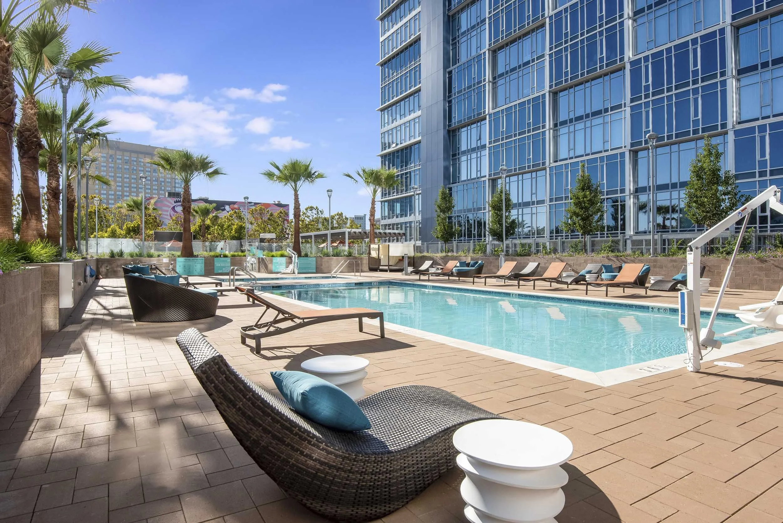 Outdoor swimming pool area with lounge chairs, palm trees, and a tall modern glass building in the background under a blue sky with some clouds.