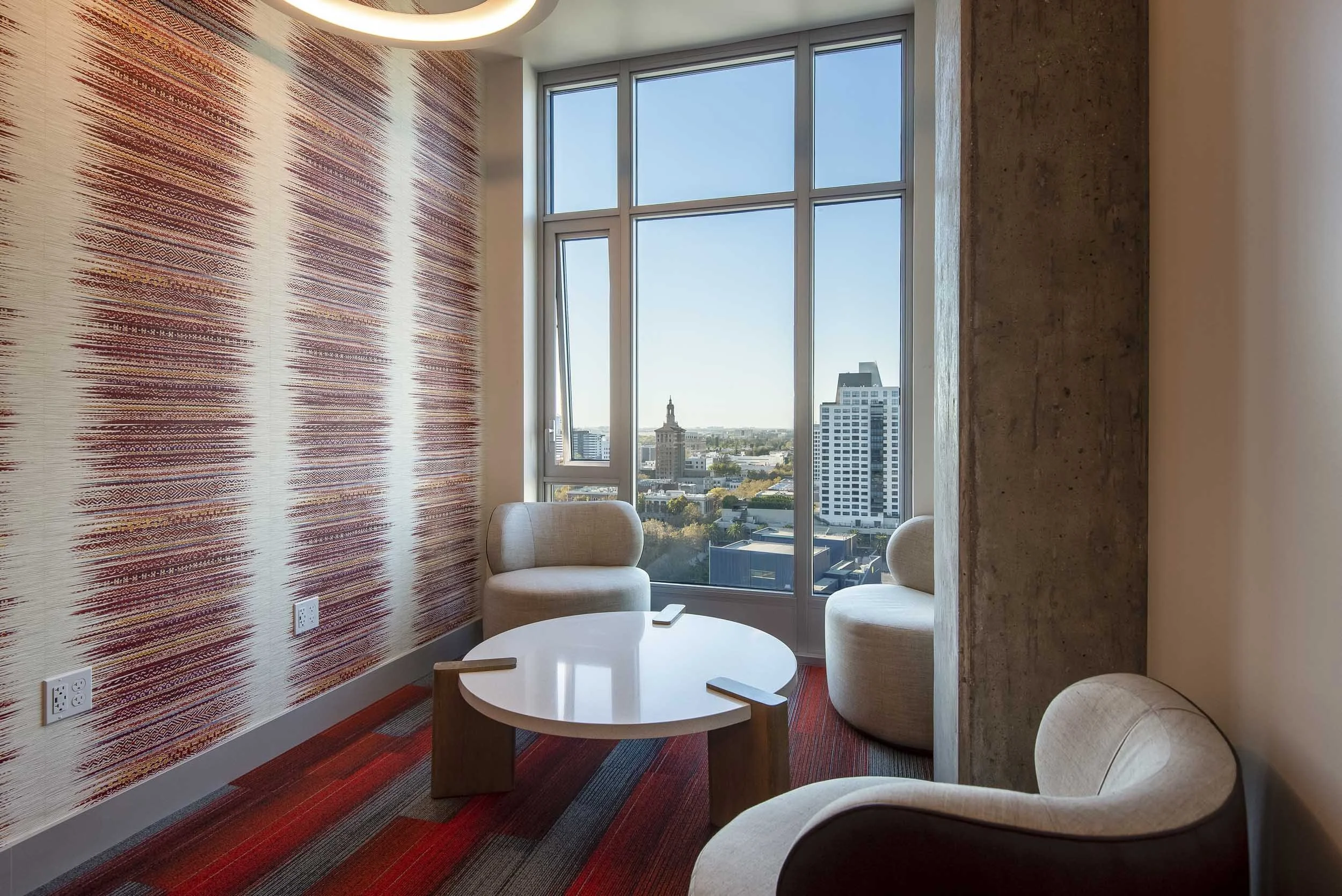 A modern interior room with a large window showcasing a city skyline, including a historic building with a clock tower. The room has a textured red and white patterned accent wall, beige chairs, a white round table, and a multicolored striped carpet.